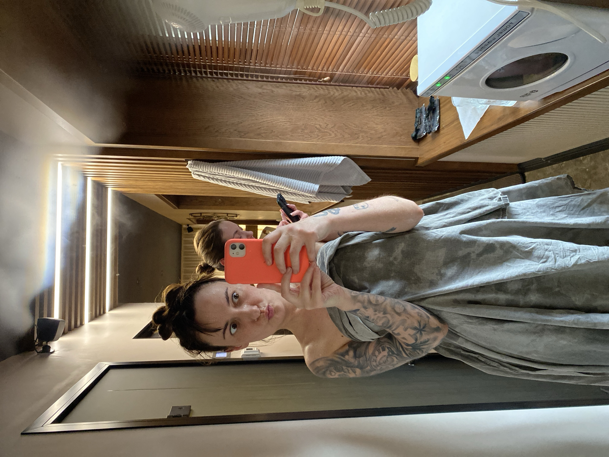 A woman with tattoos is taking a selfie in a mirror in what appears to be a spa or wellness center. She is holding an orange cell phone and is wrapped in a gray cloth. Behind her, another woman is visible holding a black cell phone. The background features modern lighting and wooden slat accents. A white appliance with a window is visible above and to the right. The location in the city of Votkinsk, Russia, cannot be determined from the image.