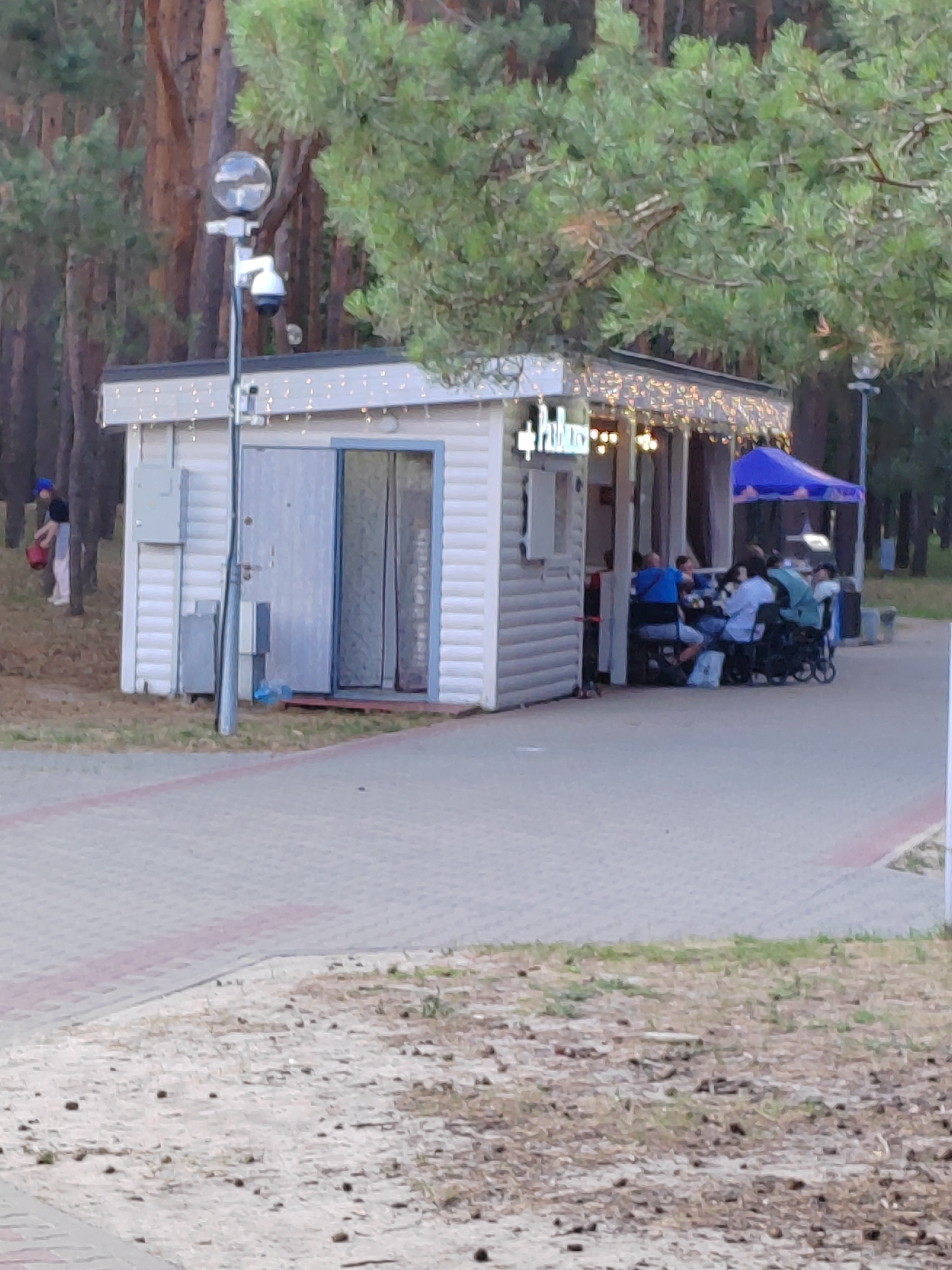 The image depicts an outdoor scene in Brest, Belarus, featuring a small white building that appears to be a kiosk or a small cafe. The building has a covered outdoor seating area decorated with string lights, where several people are seated at tables. One person is visible outside the building to the left, wearing a blue cap and holding a red bucket. A lamppost with a security camera is positioned in front of the building. The area around the building is paved with interlocking bricks, and beyond that, there is a grassy area with trees in the background, suggesting a park or wooded environment.