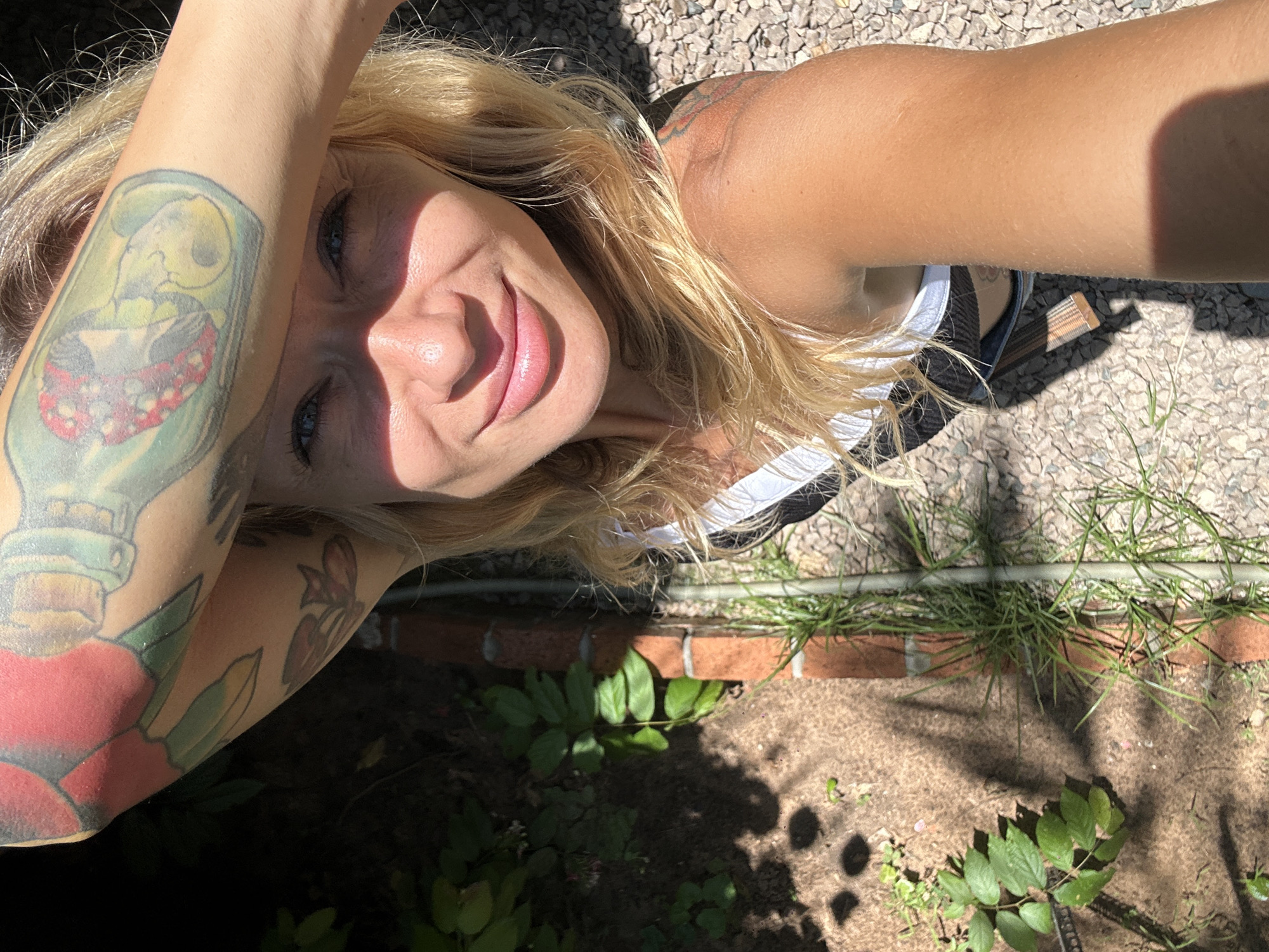 The media file is a close-up, horizontal, high-angle shot of a blonde woman lying down outdoors, with her face angled towards the camera. She is wearing a white tank top with black stripes. The woman has blue eyes and is smiling, with her left arm raised to partially cover her face, casting a shadow. Visible tattoos adorn her left arm, including what appears to be a skull within a lightbulb and a heart with red and pink petals. Her right arm is also visible, showing a tattoo on her shoulder.
The background suggests an outdoor setting with natural elements. The ground is covered in small, light-colored stones, with patches of dirt and green foliage. A brick structure is partially visible in the lower portion of the frame, bordering an area of dark soil with more plants. Sunlight creates strong shadows on the ground, indicating it is likely daytime with bright sun.
The overall mood is relaxed and cheerful, conveyed by the woman's smile and the sunny atmosphere. The location context of Mũi Né, Vietnam, suggests a warm, possibly coastal environment, although this is not explicitly depicted in the cropped image. There is no visible text in the media file.