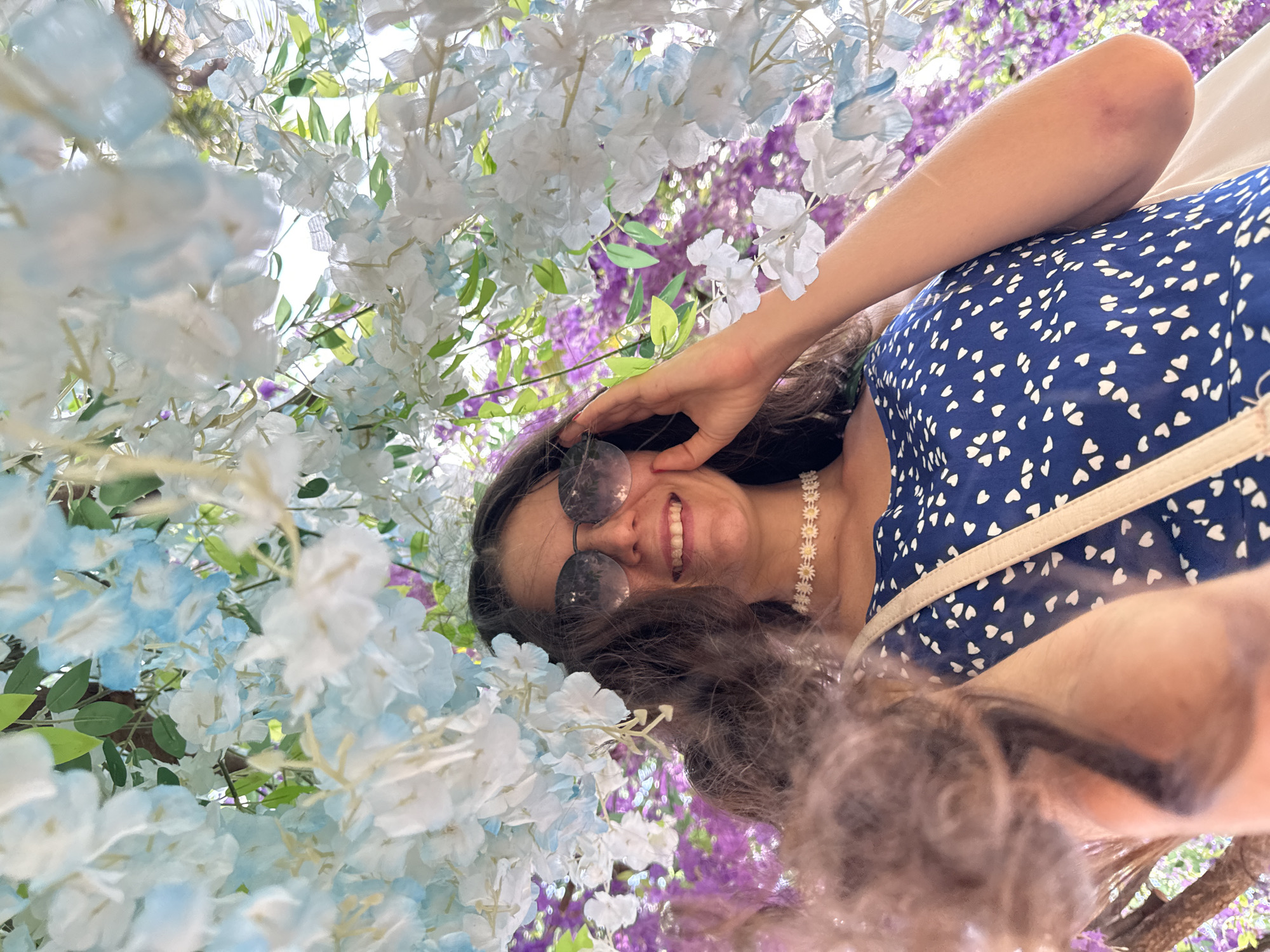 A woman is lying down and looking up at the camera, surrounded by artificial flowers. She has long, dark hair and is wearing round sunglasses, a blue dress with a white heart pattern, and a white daisy choker necklace. Her arm is bent, and her hand is resting on her forehead. She is smiling broadly, showing her teeth, and her eyes are crinkled with happiness. The scene is dominated by clusters of white and pale blue flowers, with hints of purple flowers in the background. The lighting suggests it is daytime, possibly with bright sunlight filtering through the flowers. The overall impression is one of joy and contentment, perhaps captured during a visit to a floral-themed attraction in Mũi Né, Vietnam. There is no visible text in the image.