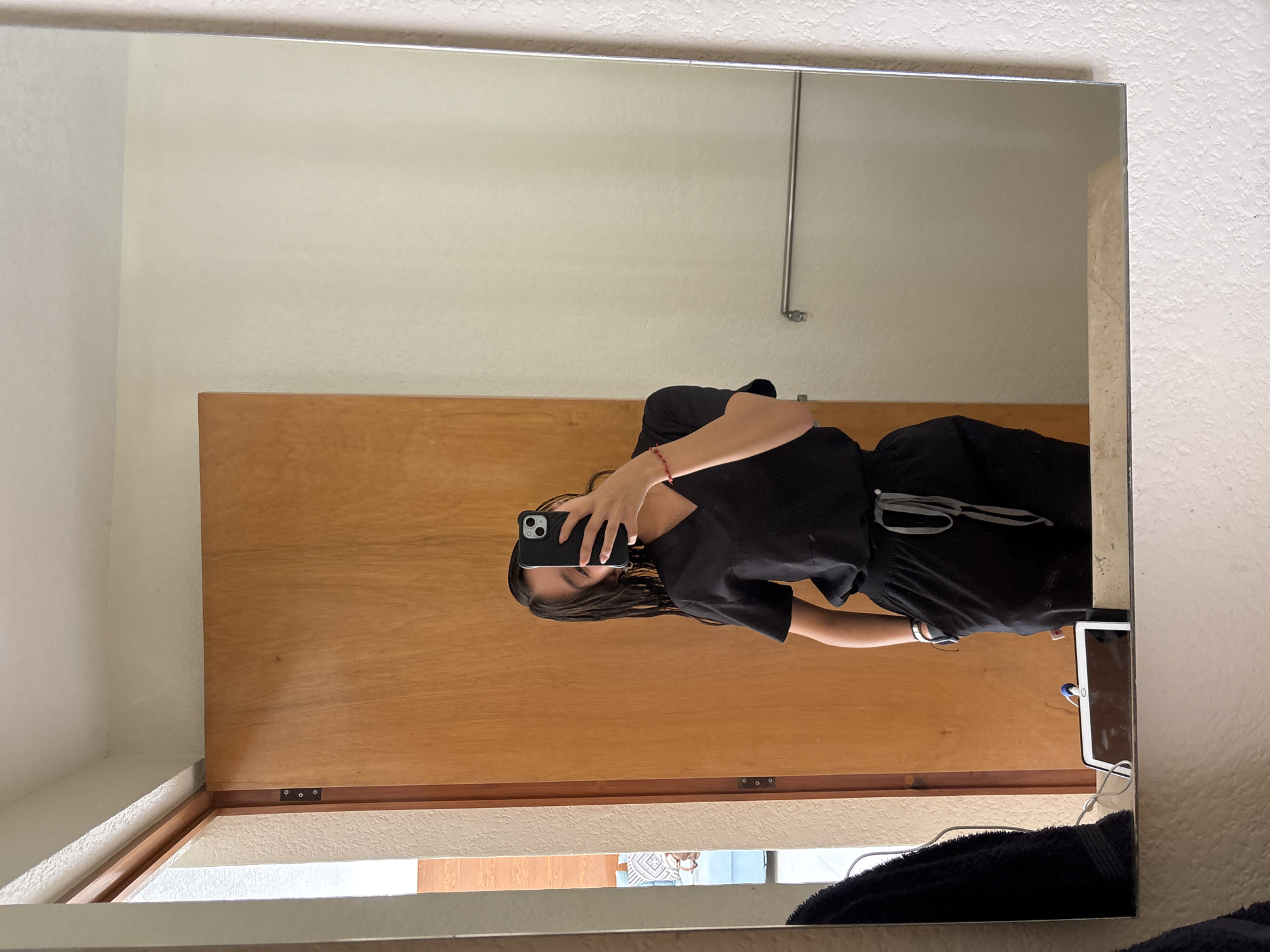 A person in a black outfit stands in front of a mirror, holding a black phone to take a selfie. The background includes a wooden cabinet and a white wall. There is a glimpse of what appears to be a window area at the bottom of the reflection. No specific information related to the city of Puebla, Mexico is apparent in the scene.