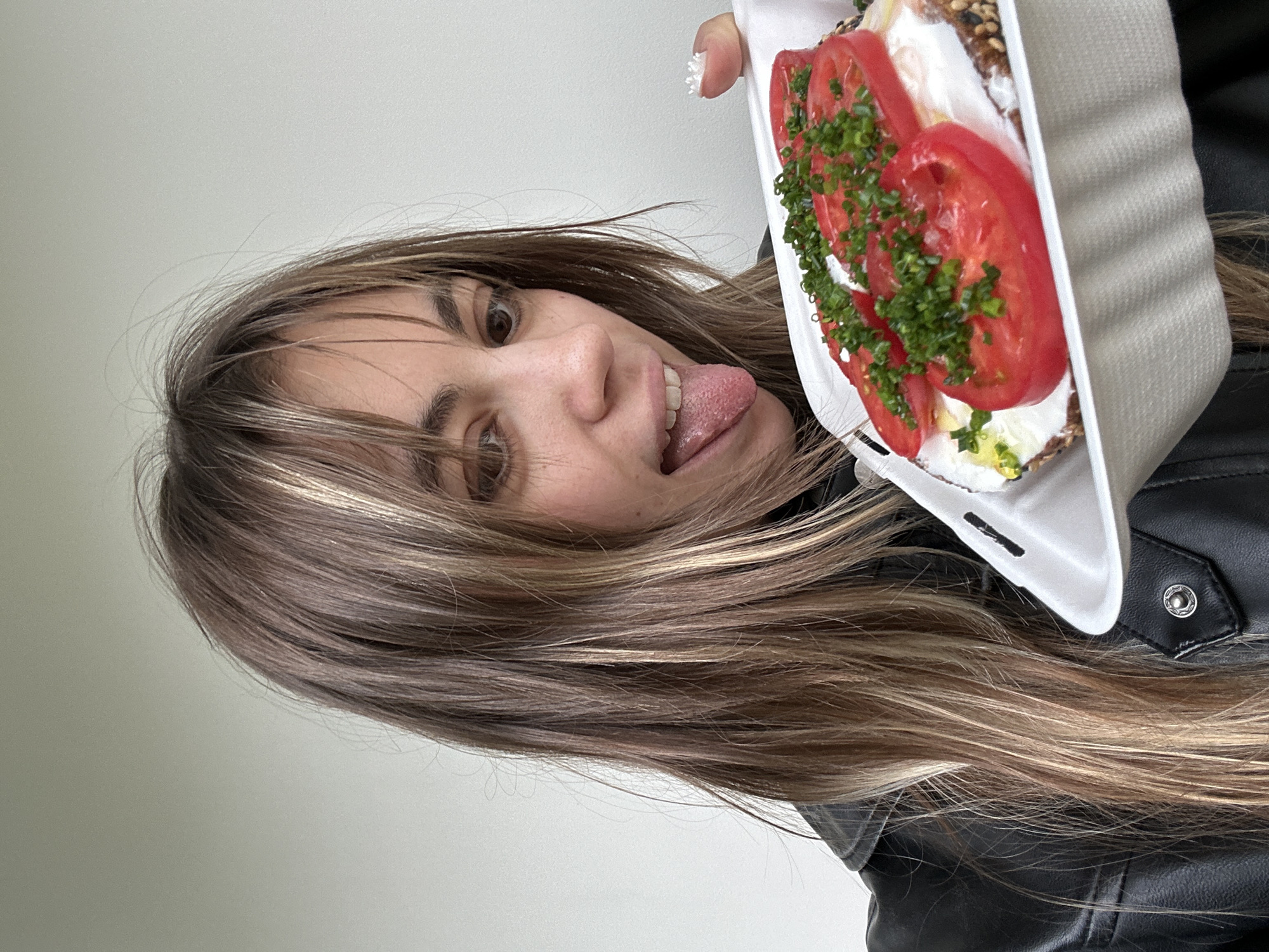 The image shows a young woman, likely in her late teens or early twenties, holding a takeaway container with food. She is looking at the camera with her tongue sticking out, which suggests a playful or mischievous mood. The food in the container appears to be some kind of open-faced sandwich or tart, with layers of cream cheese, sliced tomatoes, and chopped chives on top of a dark-colored bread base. Her hair is long and brown with lighter highlights, and it is falling over her face. She is wearing a black leather jacket. The background is a plain, light-colored wall, offering no specific clues about the location beyond a general indoor setting. The lighting suggests it could be daytime, but there are no clear indicators of the time of day or weather conditions. The image is a close-up, focusing on the woman and her food. There is no visible text in the image. The location context of Moscow, Russia, does not provide any specific visual cues in the image itself.