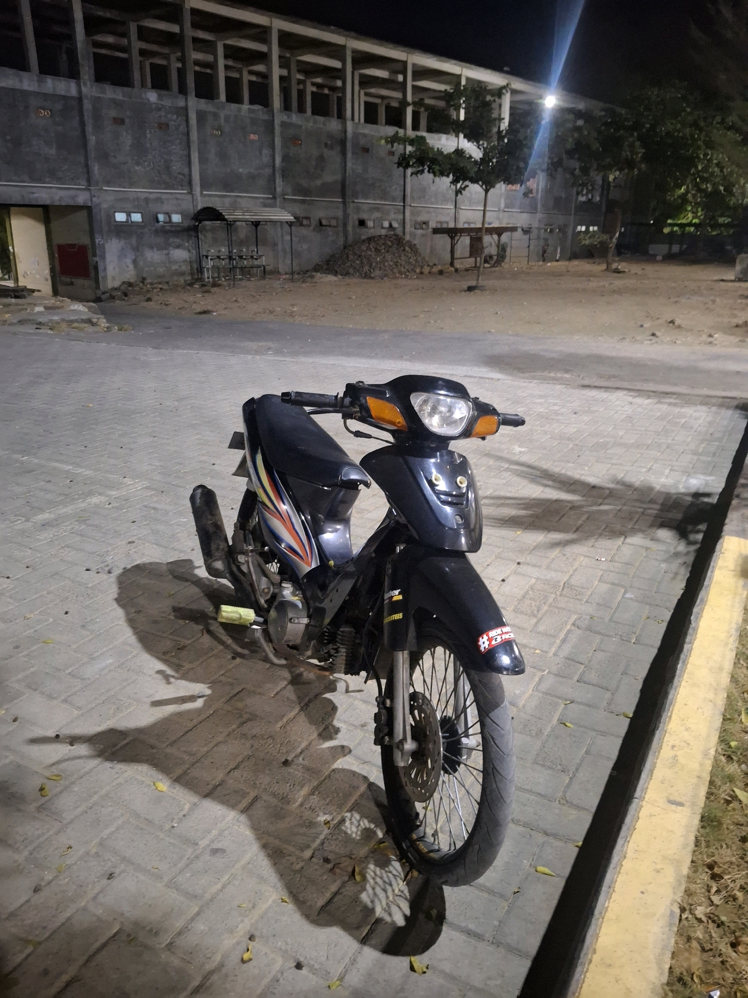 An outdoor, nighttime, medium shot shows a dark blue and black motorcycle parked on a paved walkway in Potoan Laok, Indonesia. The motorcycle is positioned facing the viewer's left, with its front wheel slightly turned. The bike has a black seat with colorful graphics on the side. The front of the motorcycle features a headlight and orange turn signals. A black exhaust pipe is visible on the left side. The walkway is made of light brown pavers. In the background, there is a large, unfinished concrete building with columns and openings. To the right of the building, trees are visible. The ground behind the walkway appears to be dirt with some piles of debris. A yellow line runs along the edge of the walkway on the right side of the frame. The lighting suggests artificial light sources, possibly streetlights, casting shadows from the motorcycle and surrounding objects onto the paved surface. The scene appears to be a public area, possibly a parking lot or an open space adjacent to the building.