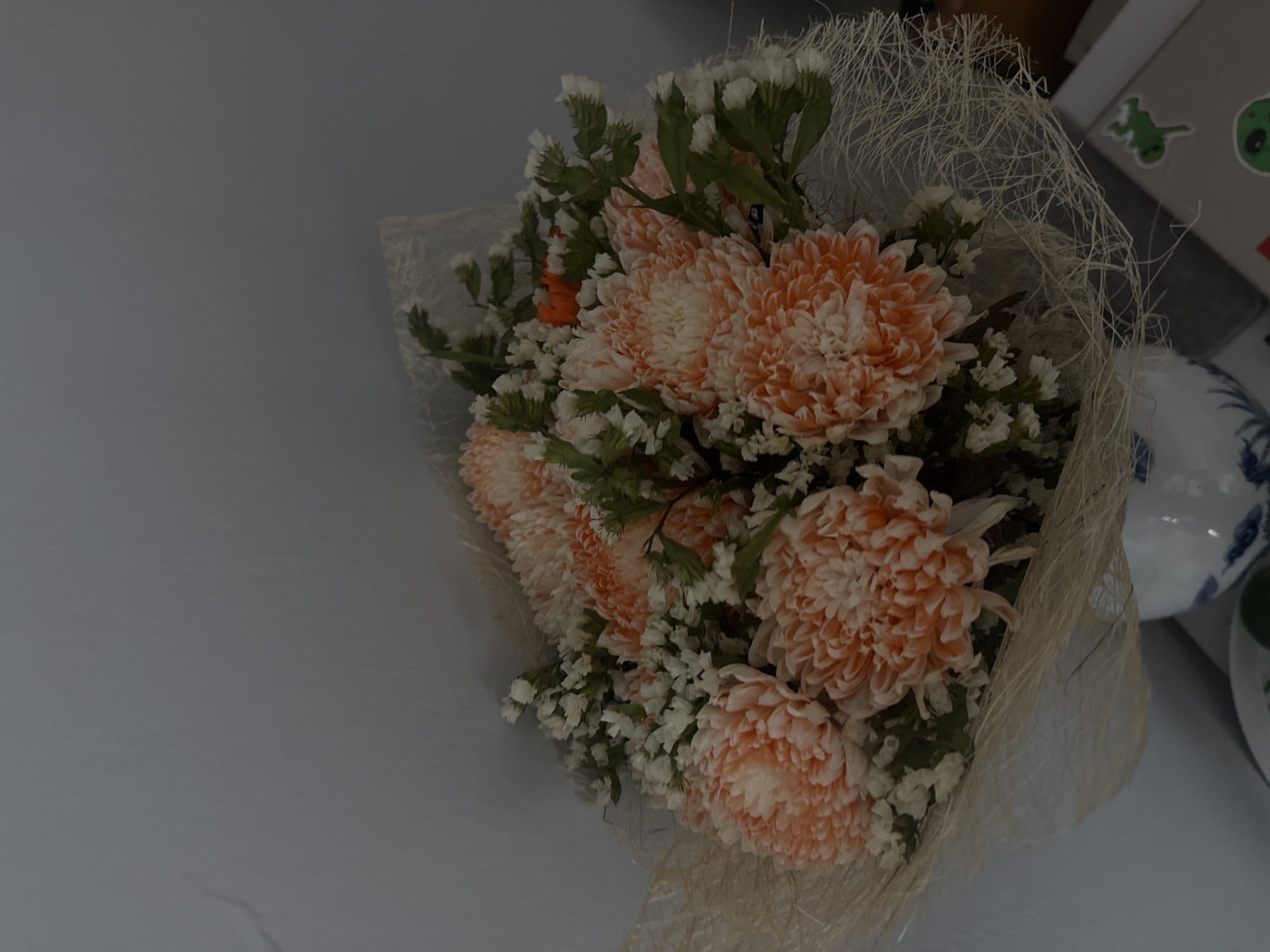 A bouquet of orange chrysanthemums and white filler flowers is displayed in a white vase with blue floral patterns. The bouquet is wrapped in a textured, light-colored material. The background is a plain, light gray wall. The scene appears to be indoors, possibly in a home or office setting in Nha Trang, Vietnam. There are no people visible in the image.