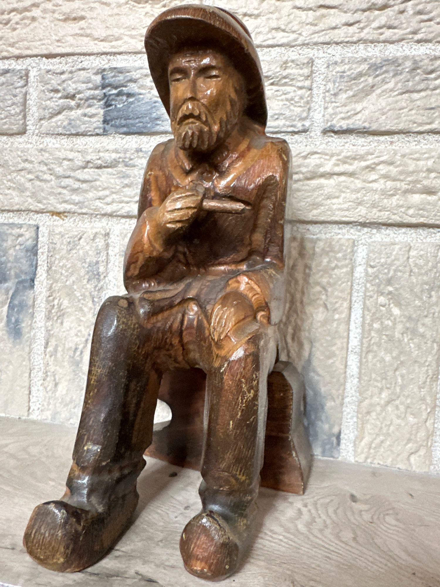 A wooden carving of a man sitting on a stool is positioned in front of a textured brick wall. The carving depicts a man with a hat, beard, and detailed clothing, holding something in his hands. The background is a light-colored brick wall with horizontal and vertical grout lines, and some areas show a bluish-gray discoloration. The carving is placed on a wooden surface with a visible wood grain pattern. The scene appears to be indoors, possibly a domestic setting or a craft display. The specific location in Пінкавіцкі сельскі Савет, Belarus, cannot be determined from the image alone.
