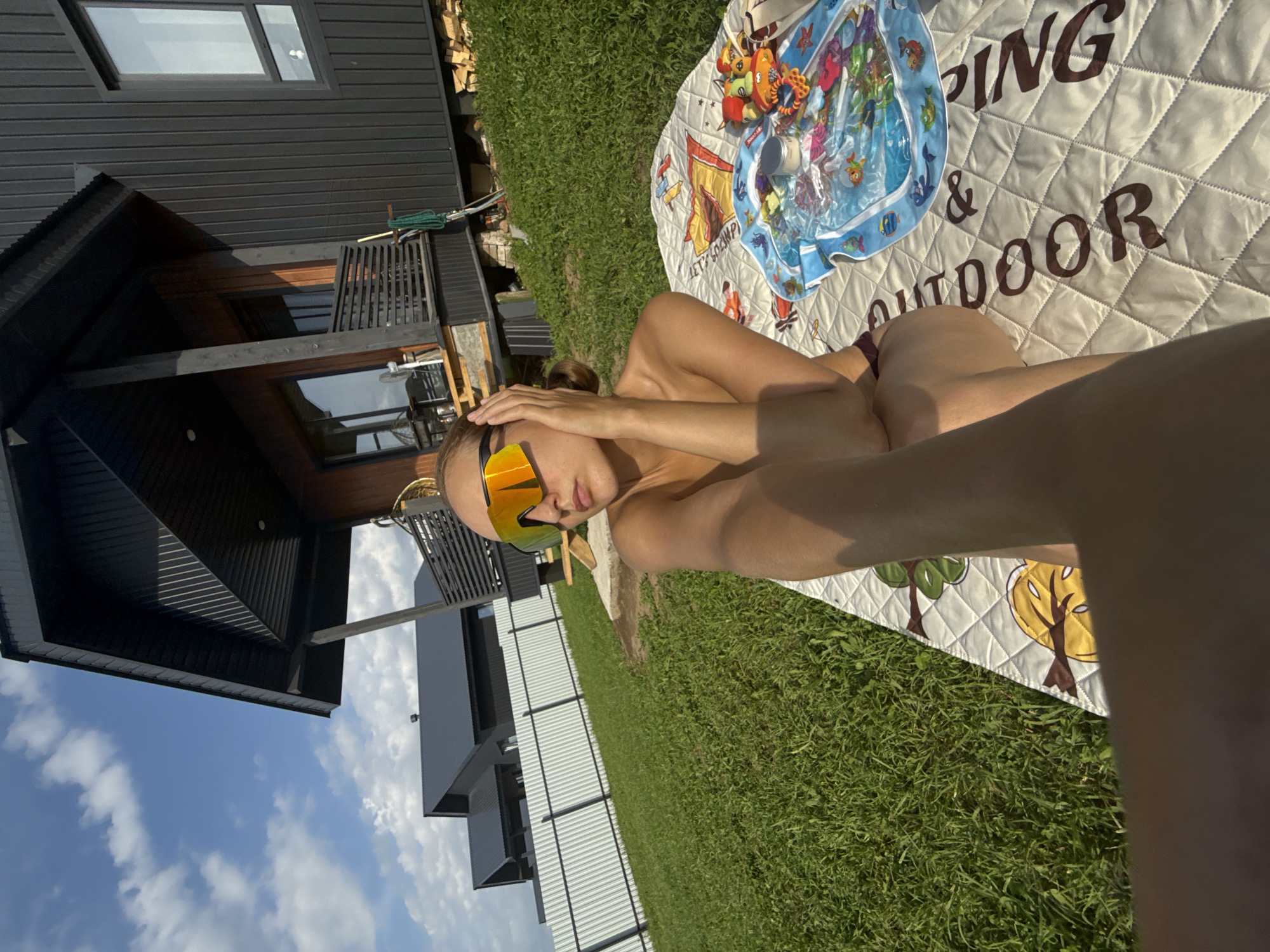 A person is lying on their back on a quilted blanket on a patch of green grass. The person is wearing large, reflective sunglasses with orange and yellow lenses. They are holding one hand up to their forehead, obscuring part of their face. The blanket has a quilted pattern and features cartoonish illustrations of trees and other motifs. A blue and white inflatable mat with colorful aquatic imagery and some text is also on the blanket.
In the background, to the left, is a modern, dark-colored house with several large windows. A wooden deck or balcony area is visible. Beyond the house, there is a glimpse of a blue sky with scattered white clouds, suggesting it is daytime and the weather is likely fair. To the right of the person, stacks of firewood are visible. The overall scene appears to be set outdoors, possibly in a backyard or recreational area. The text on the blanket reads "CAMPING & OUTDOOR". The image is oriented sideways.