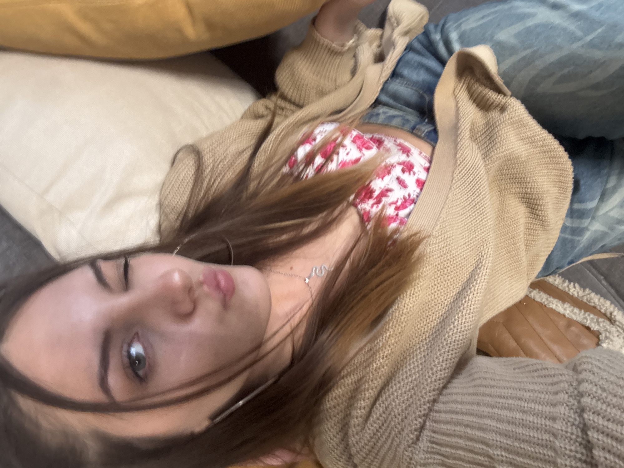 A young woman is lying on a gray sofa. She is wearing blue jeans, a white top with red floral print, and a beige cardigan. She is lying between two pillows, one yellow and one beige. She is wearing silver hoop earrings and a silver necklace with a snake pendant. She is looking at the camera and winking. Her hair is brown. The location cannot be determined from the image.
