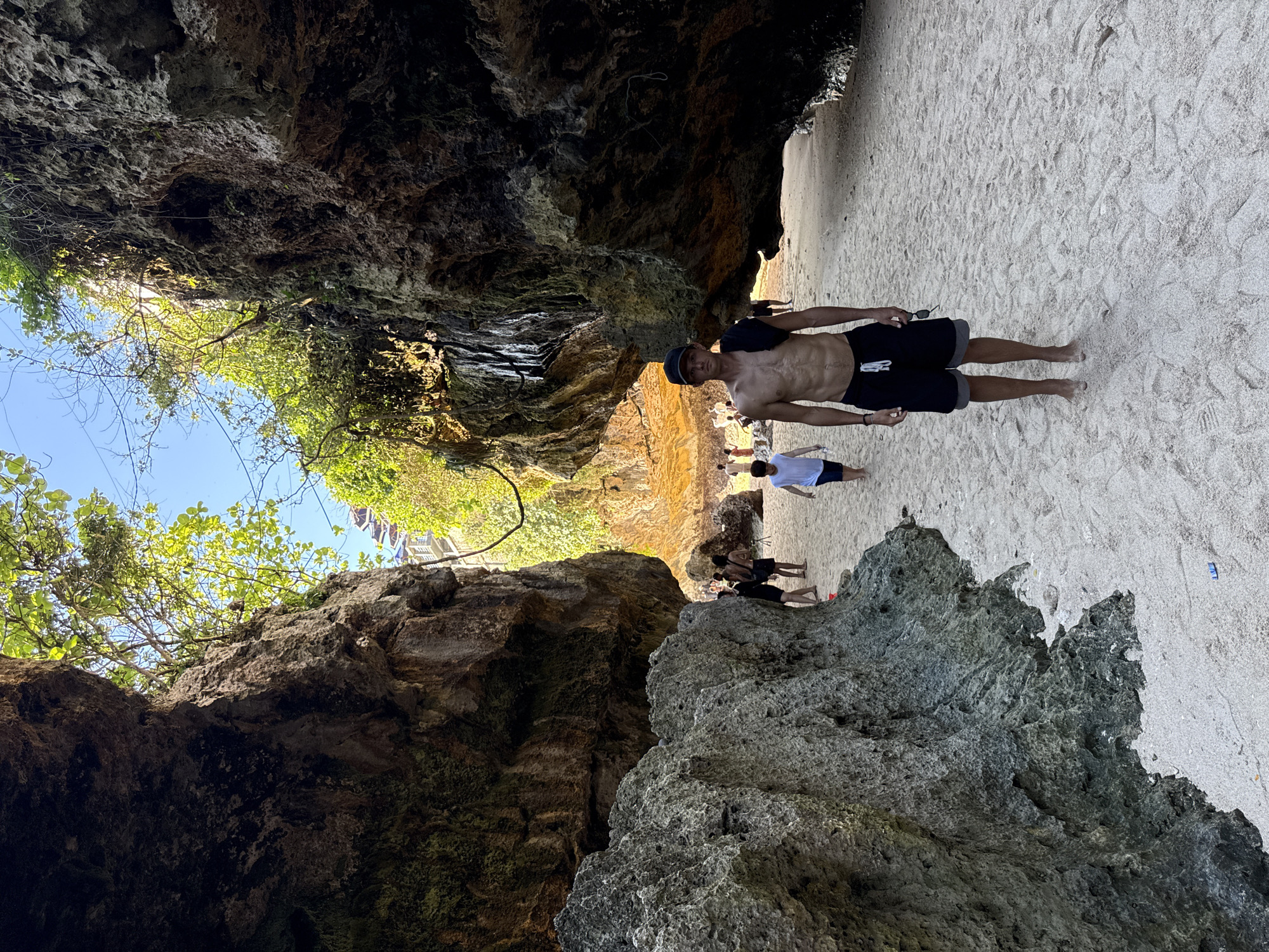 This image captures a vibrant coastal scene in Kuta Selatan, Indonesia, on a clear, sunny day. The primary focus is a natural passage or cove, formed by towering, rugged rock formations that create a shaded pathway.
In the foreground, a shirtless man wearing dark shorts and a dark baseball cap stands on the sandy ground, looking directly at the camera. He holds a pair of sunglasses in his left hand. The sand underfoot is light-colored and fine, with visible footprints.
Beyond the man, several other individuals are present, walking or standing further down the passage. One person in a light blue t-shirt and dark shorts is seen walking away, while others are less distinct in the middle distance.
The rock formations on either side are imposing, displaying varied textures and shades from dark, shadowy crevices to lighter, sunlit surfaces. Above these rocky walls, a brilliant blue sky is visible, framed by lush green foliage, including trees and bushes growing on the cliffs and reaching towards the sunlight. The bright lighting suggests it is daytime, likely mid-morning or early afternoon. In the background, beyond the immediate rock passage, hints of buildings or structures can be discerned through the greenery, suggesting human development near this natural landscape. The overall scene depicts a leisurely activity, possibly exploring a unique beach or coastal pathway. There is no visible text in the image.