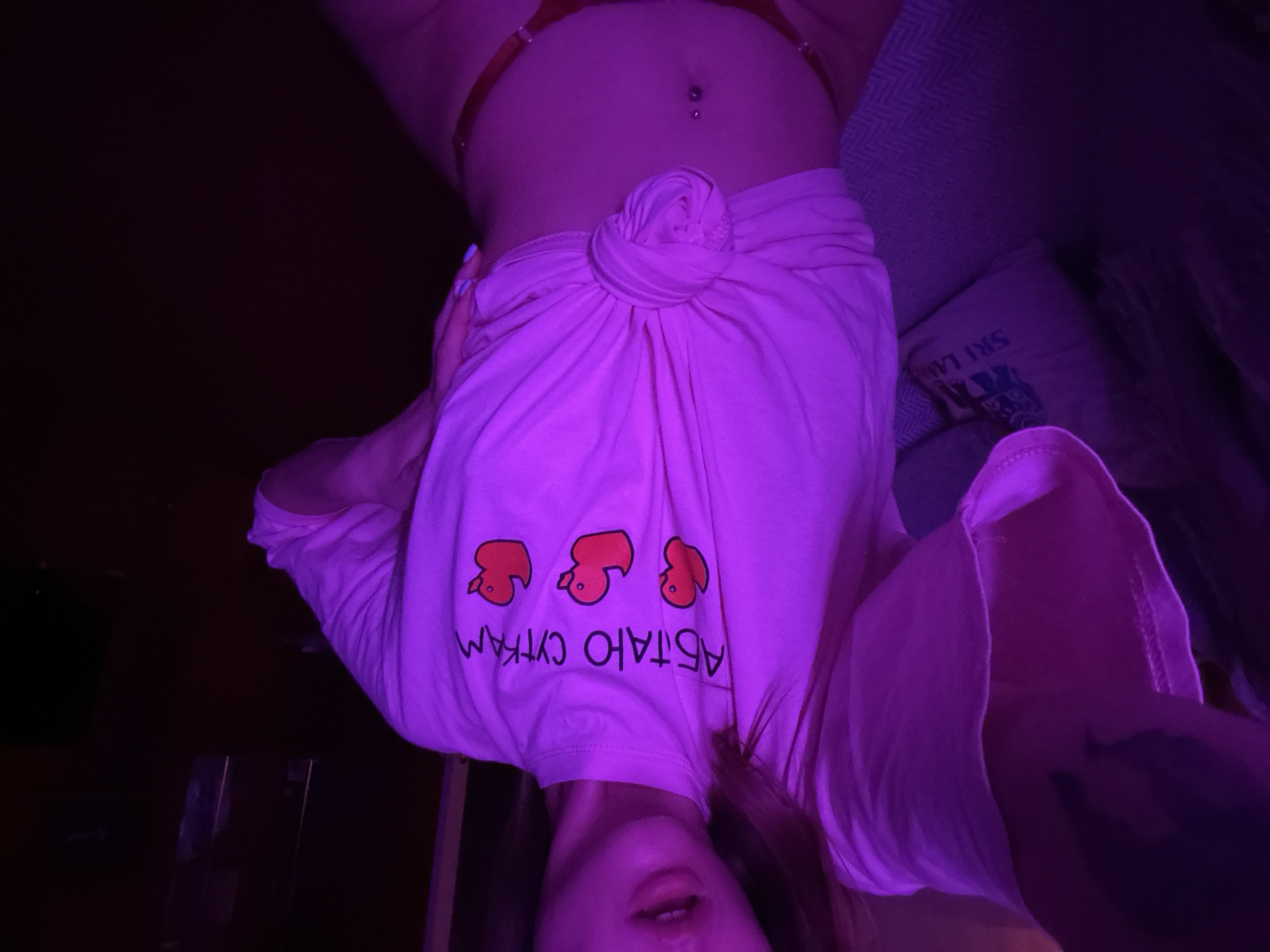 The image depicts a person, whose face is partially visible, upside down in a dimly lit room with purple lighting. The subject is wearing a light pink t-shirt with a knot tied in the front, adorned with three orange duck-like figures and Cyrillic text. The text reads "АБТАЮ СУТКАМ," which translates to "I melt away the days." Beneath the t-shirt, a portion of what appears to be an orange undergarment is visible. The person also has a belly button piercing.
In the background, a pillow with the words "SKI LAB" and an illustration of a ski resort is visible, suggesting a potential connection to a winter vacation or theme. The setting appears to be indoors, possibly a bedroom or living space. The overall mood is intimate and playful, enhanced by the low lighting and unusual orientation of the subject. The time of day cannot be determined, and no weather cues are present. The location context is given as Khimki, Russia.