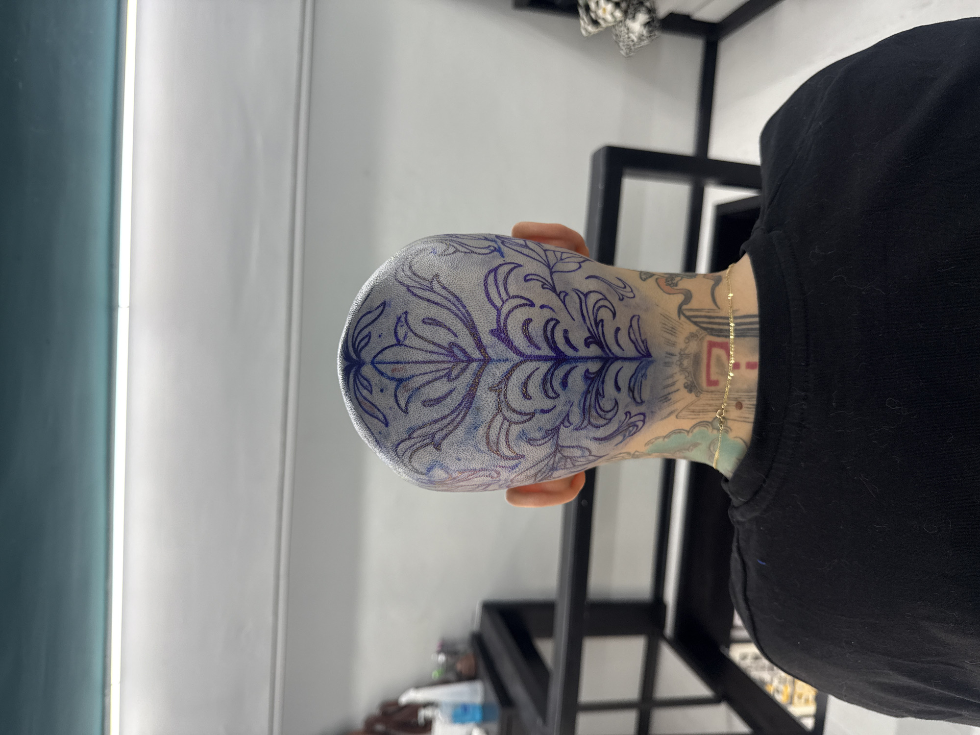 The image is an overhead shot of a person's head with a detailed tattoo design. The person appears to be wearing a black shirt, and a gold chain is visible around their neck. The tattoo on the head is intricate, featuring a botanical or floral-inspired pattern in shades of blue and possibly purple. Other tattoos are visible on the neck and upper back. The environment includes a white wall with linear accents and parts of a black metal structure. The image does not provide sufficient detail to determine a location in Sofia, Bulgaria.