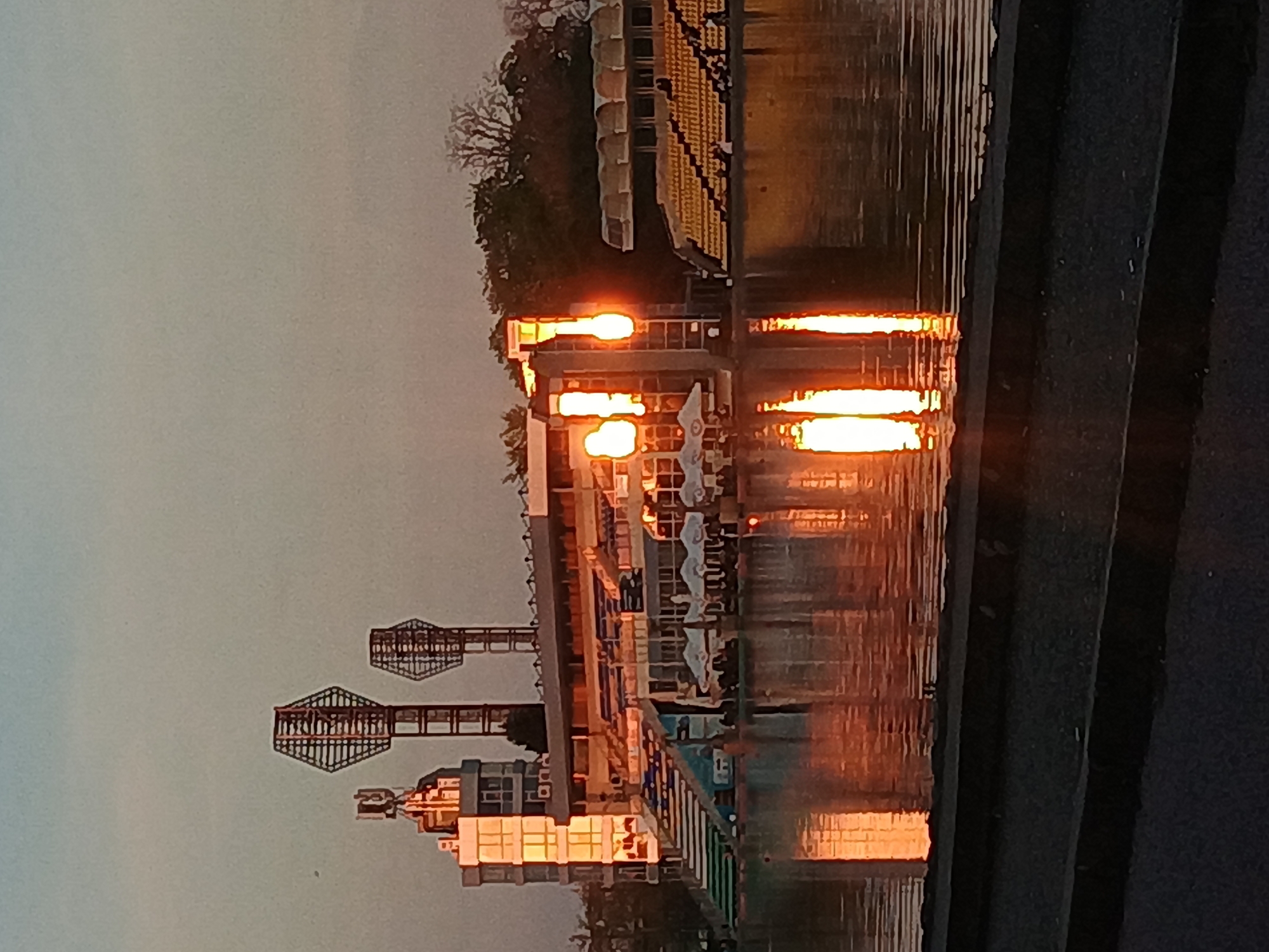 This media file captures a twilight or nighttime scene of a waterfront area, likely a rowing canal or river, lined with buildings and structures. The image is originally oriented vertically but depicts a landscape view.
In the mid-ground, a wide body of water reflects the bright artificial lights from the structures along its far bank, creating prominent vertical streaks of orange and yellow light on the water's surface. On the left side of the far bank, a stadium with numerous empty yellow seats is visible, partially obscured by trees. Below the stadium, a building with several intense lights illuminating its exterior casts strong reflections. Further to the left along the bank, two tall, intricate lattice-work towers, possibly floodlight pylons or architectural features, stand against the dim sky. Adjacent to these, another multi-story building with warmly lit windows is discernible. Several small boats or sculls are docked along the bank near the main lighted building. The sky above is a muted grey, suggesting either late evening or early morning. In the immediate foreground on the right, a dark, textured surface, possibly a concrete embankment or pier, frames the scene.
The strong, warm glow of the artificial lights against the dark sky and water creates a serene yet dramatic atmosphere. No people or specific activities are visible, implying a quiet moment. Given the location context of Plovdiv, Bulgaria, this scene likely depicts a portion of the Plovdiv Regatta Venue, known for its rowing course and stadium. There is no visible text in the image.