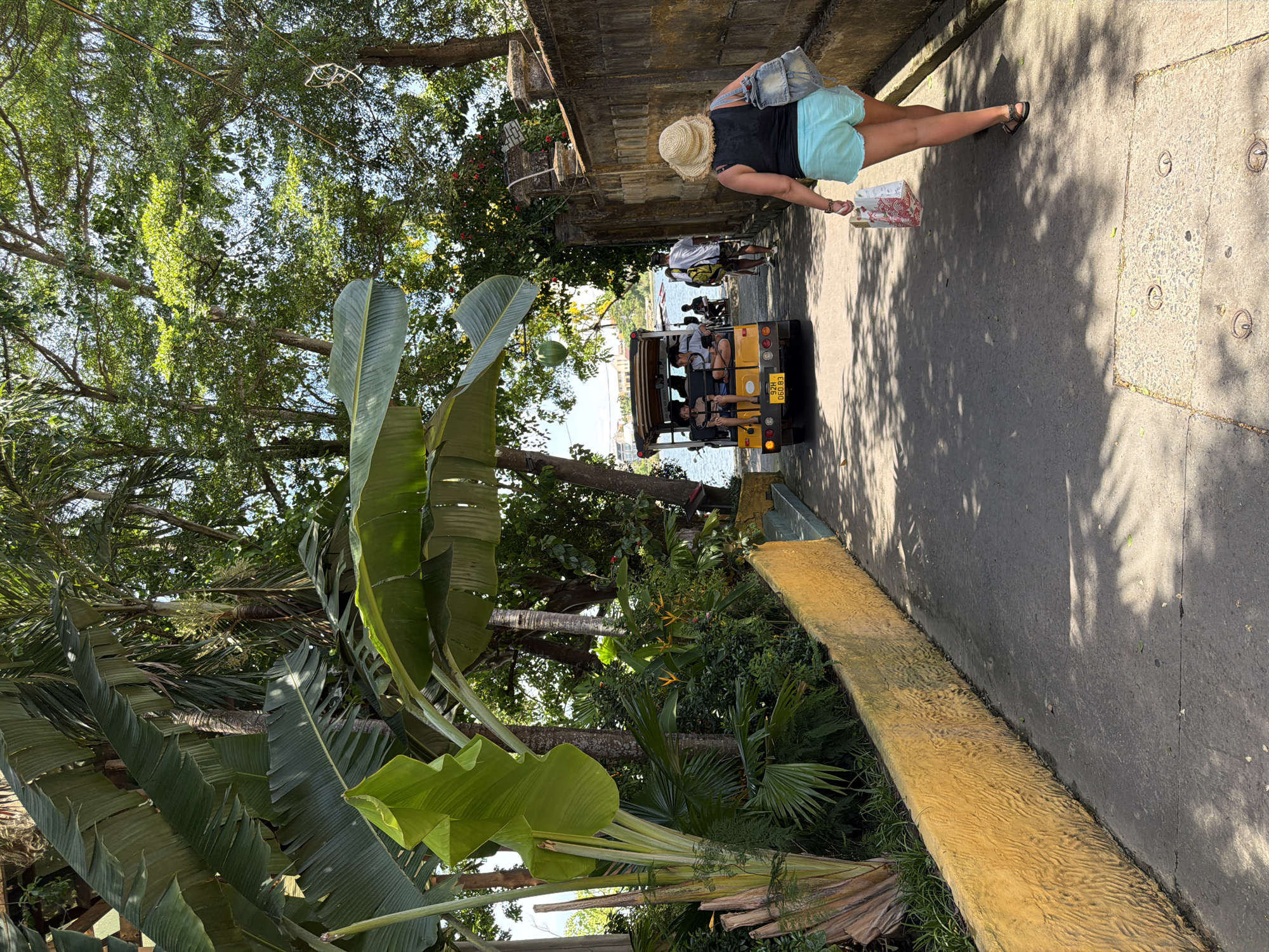 An eye-level, medium shot captures a paved walkway in Hội An Ward, Vietnam, bordered on the left by a textured, bright yellow retaining wall and lush tropical foliage, including large banana leaves and palm trees. On the right, a weathered, stone wall runs along the path. The sun casts dappled shadows across the walkway. In the foreground, a person wearing a straw hat, a black tank top, and light blue shorts walks away from the camera, carrying a patterned shopping bag and a denim bag slung over their shoulder. Further down the path, a yellow open-air electric vehicle, similar to a golf cart, carries several passengers. A man wearing a white t-shirt and shorts walks ahead of the vehicle. In the background, a body of water, likely a river or canal, is visible, with buildings and more greenery on the far bank. The scene appears to be a picturesque and tranquil area within Hội An.