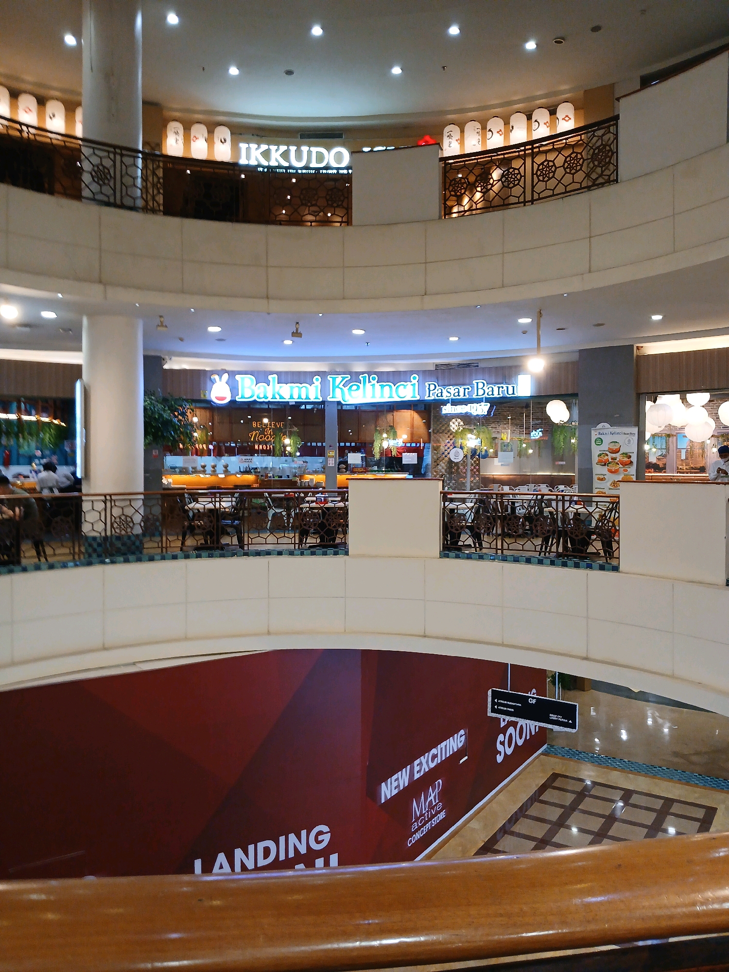 This is an indoor shot of a multi-level shopping mall, likely in North Jakarta, Indonesia. The scene features two levels visible, with a central atrium.
On the upper level, a restaurant called "IKKUDO" is visible, identified by its signage and decorative paper lanterns. Further along, another establishment with the signage "Bakmi Kelinci Pasar Baru since 1957" is also present, with an open kitchen and seating area.
The middle level shows a railing with a decorative metal pattern, separating the levels and providing a view into the atrium. More seating and tables are visible on this level, belonging to the restaurants.
The lower level of the atrium features a large maroon banner with white text that reads "NEW EXCITING MAP ACTIVE CONCEPT STORE" and "LANDING SOON." A signpost indicates "GF" and directional arrows. The floor is tiled with a grid pattern and reflective surfaces from overhead lighting.
The environment appears clean and well-lit, typical of a modern shopping mall. There are no discernible people in the immediate foreground, but there are glimpses of individuals in the background within the restaurant areas and potentially walking on the lower levels. The overall atmosphere is commercial and inviting.