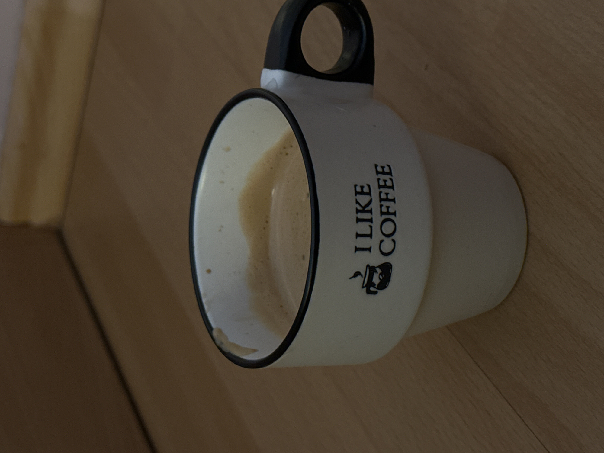 A white ceramic coffee cup, approximately 10 cm in height, with a black handle and a black rim, is positioned on a light brown wooden floor. The cup features the phrase "I LIKE COFFEE" printed in black capital letters, accompanied by a black graphic resembling a steaming coffee brewer. The cup contains a light brown liquid with a visible layer of tan-colored crema or foam, indicative of coffee. The liquid level is approximately 2 cm below the cup's rim. The wooden surface displays visible grain patterns and seams, suggesting plank construction. In the background, a darker brown surface is visible, meeting the wooden floor at an acute angle, indicative of a wall or another furniture piece. The scene is illuminated by ambient light, creating soft shadows. The location is the city of Stara Zagora, Bulgaria. No individuals or actions are depicted.