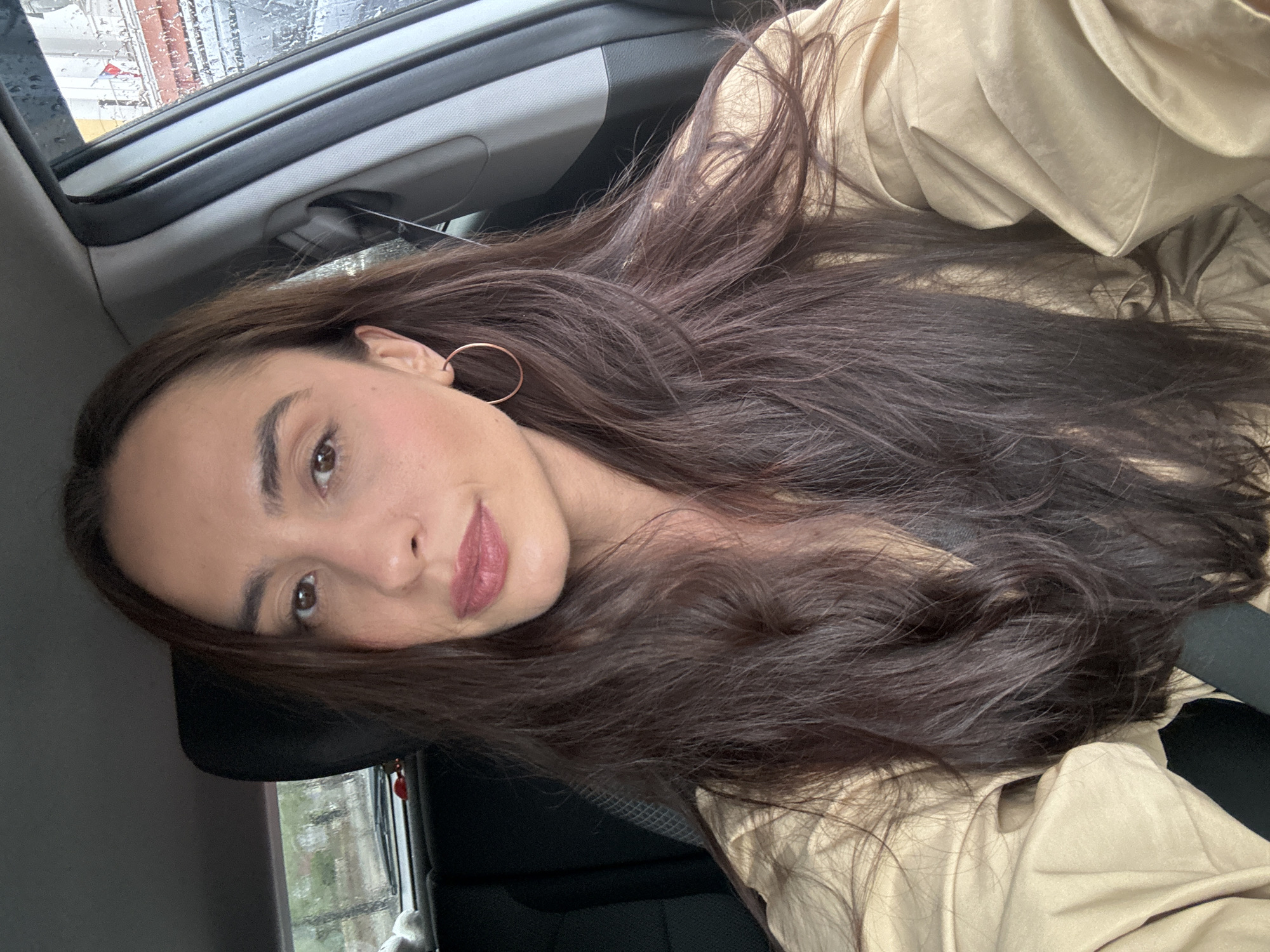 A woman with long, dark brown hair, wearing a beige trench coat and a hoop earring in her left ear, is inside a car. Rain is visible on the car's sunroof and side window. The location is possibly Krasnoyarsk, Russia.