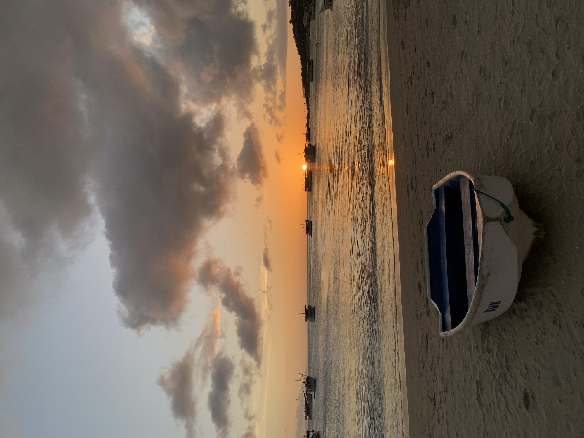 The image depicts a sunset scene at the beach in Juan Griego, Venezuela. In the foreground, a small, white boat with blue accents rests on the sandy shore. The water is calm, with gentle waves lapping at the beach. Several fishing boats are anchored in the distance, silhouetted against the vibrant orange and yellow hues of the setting sun. The sky is filled with dramatic clouds, some tinged with pink and orange from the fading sunlight. The overall atmosphere is serene and peaceful, evoking a sense of tranquility as day turns into night.