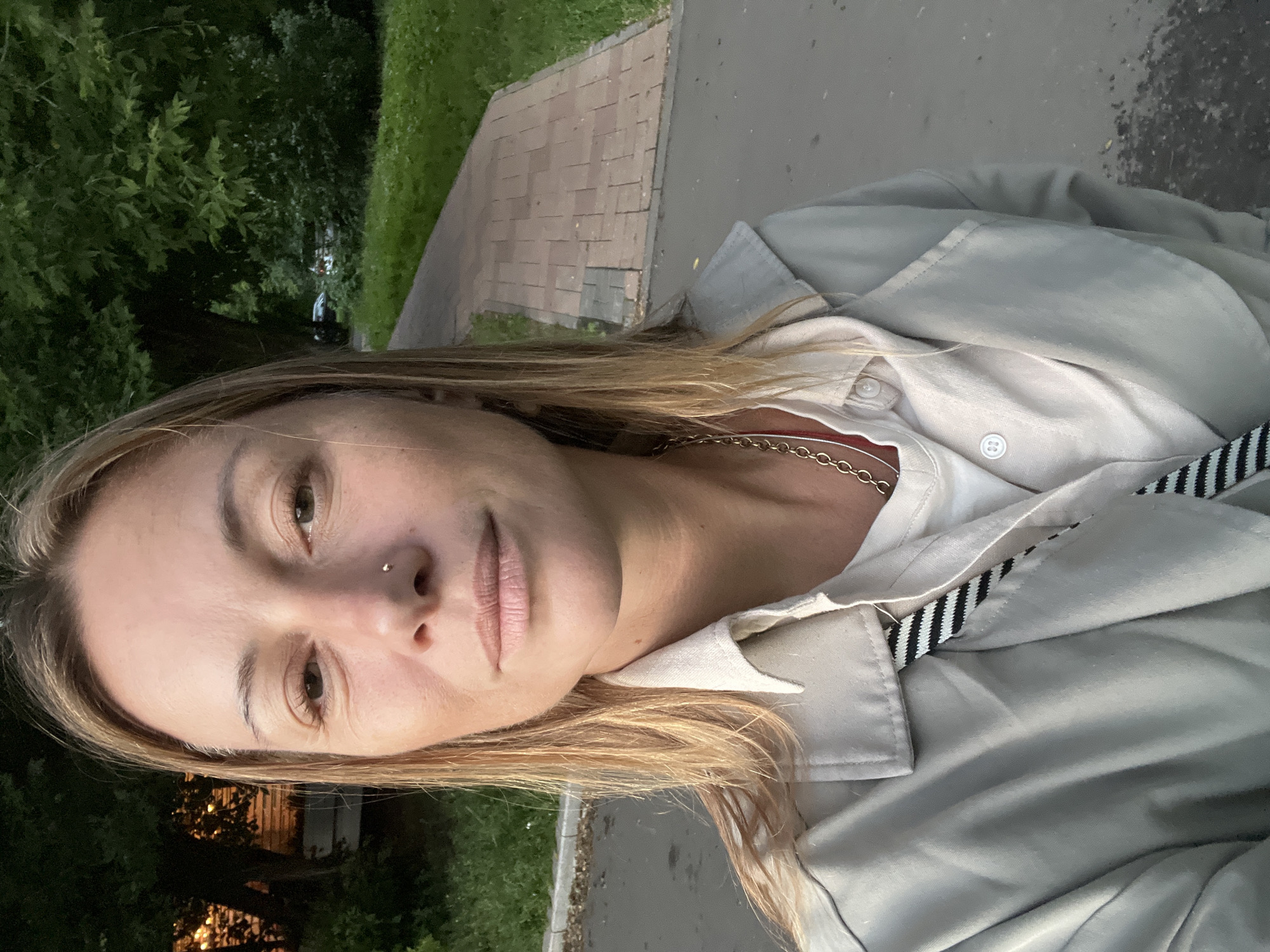 The media file is a portrait of a woman taken outdoors. She is looking directly at the camera with a neutral expression. Her hair is long and blonde, falling around her face. She is wearing a light-colored shirt and a beige jacket. A gold chain necklace is visible around her neck. A strap with black and white stripes is visible on her jacket. The background suggests an urban or suburban setting, with a paved pathway and greenery. The lighting indicates it is likely evening or dusk. The overall impression is casual and candid. There is no visible text in the image. The location context is Moscow, Russia.