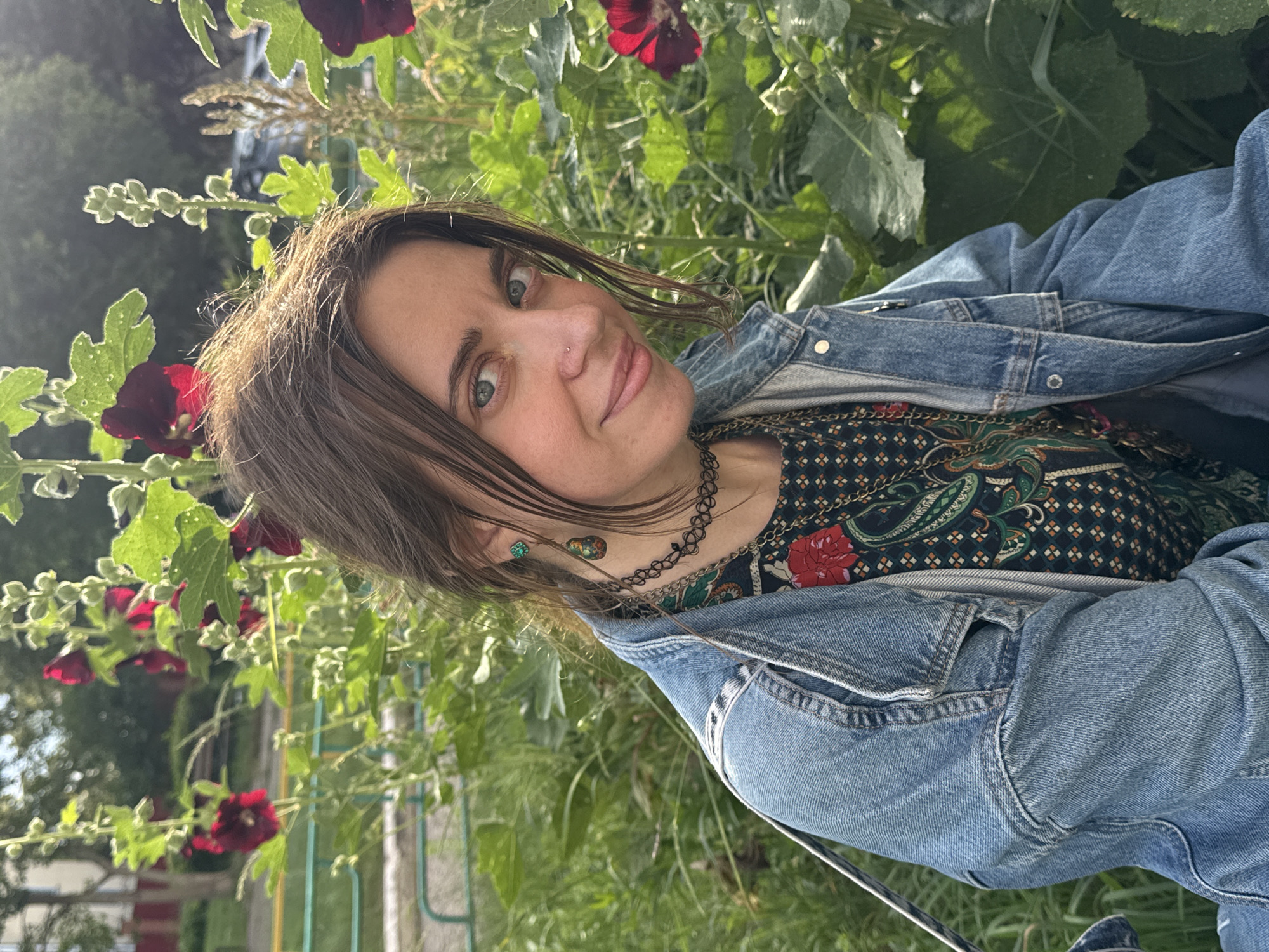 The image shows a woman outdoors, with her head and shoulders visible. She is wearing a denim jacket over a dark patterned shirt with red floral accents. A black choker necklace is around her neck, and she has a small nose stud and earrings. Her hair is dark brown and falls around her face. She is looking towards the camera with a slight smile.
The background is filled with lush green foliage and some dark red flowers, suggesting a garden or natural setting. Sunlight filters through the leaves, creating a bright and somewhat dappled lighting effect. The overall impression is of a casual, sunny day. The image is oriented sideways. No text is visible in the image.