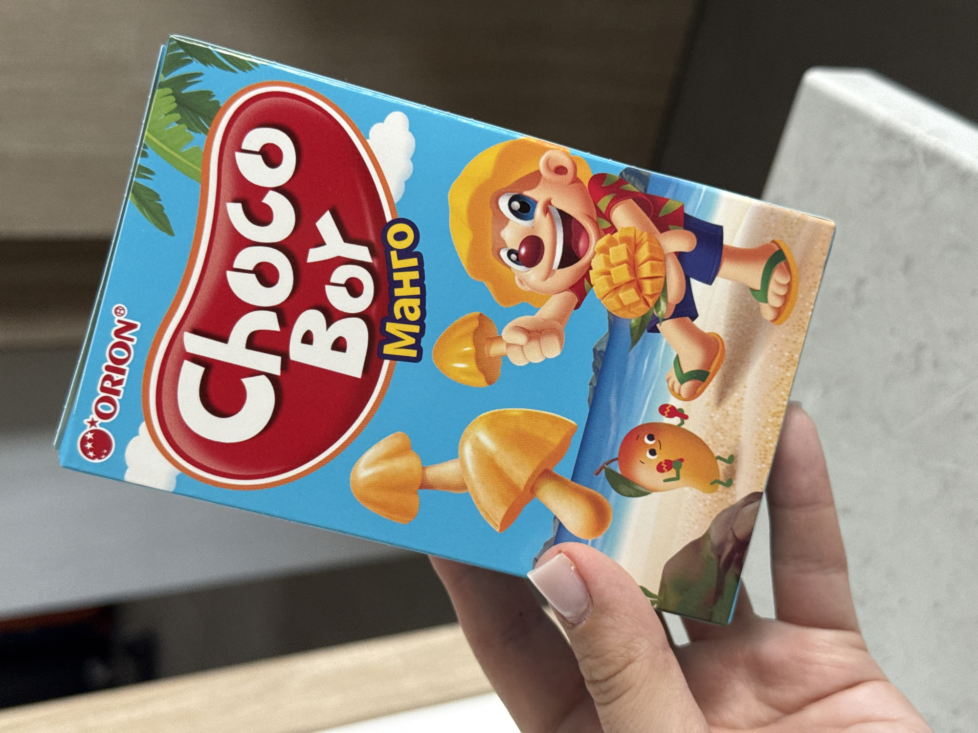 An overhead, close-up shot shows a hand holding a brightly colored juice box. The box is blue and features illustrations of a beach scene with cartoon characters. A character, seemingly a young boy with a yellow hat and red shirt, sits on the sand holding a mango. Other elements include cartoonish mushrooms and a mango character. The box has text in English and Russian, including "Choco Boy" and "Манго" (Mango). The hand holding the box has a light skin tone and a neatly manicured fingernail. The background is blurred, but appears to be an indoor setting with neutral tones. There is no discernible connection to Lyubertsy, Russia.