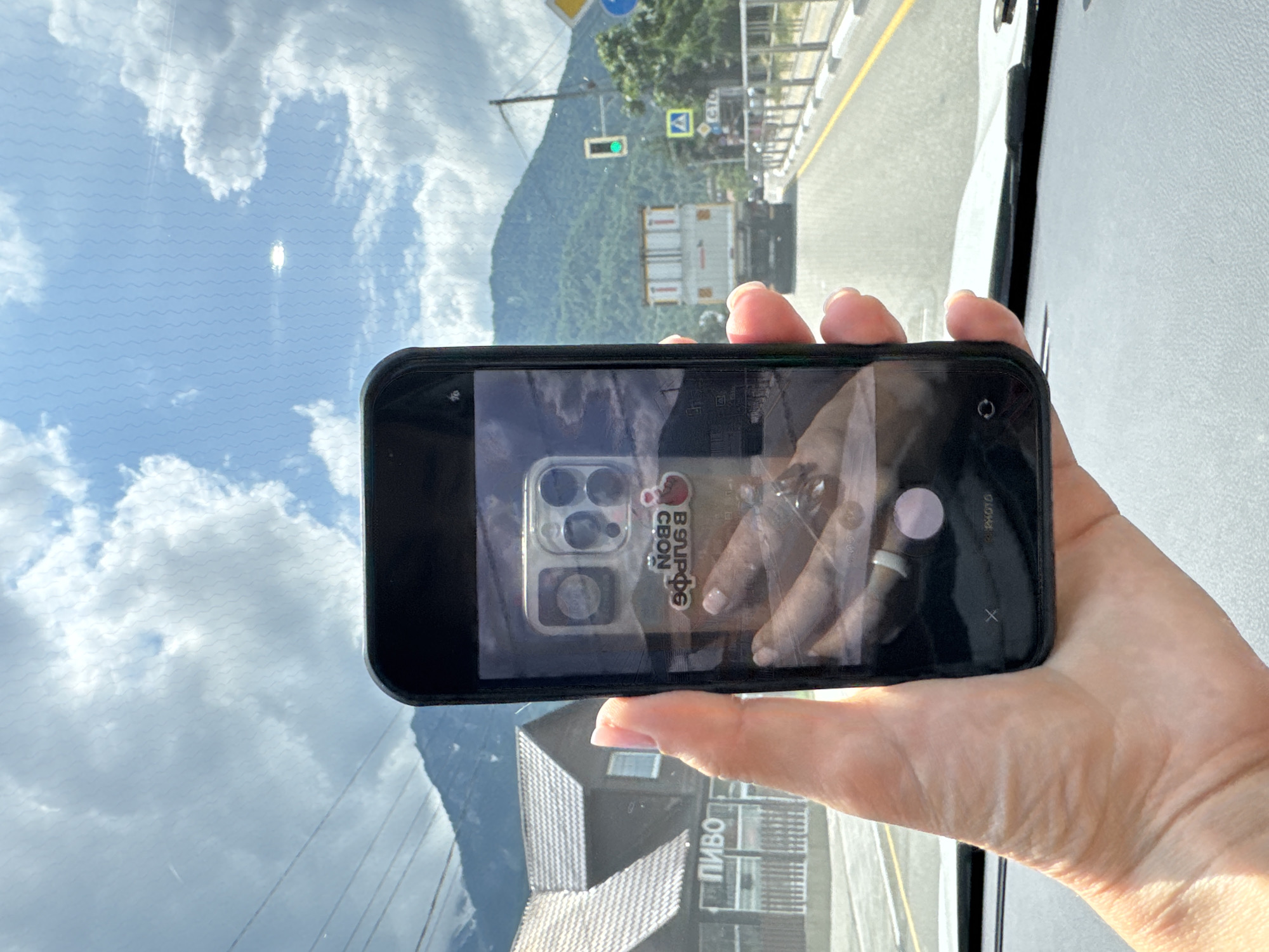 A person's hand holds a smartphone to take a picture of a street scene. The phone's screen displays a reflection of the person's hand and the phone's camera. The background visible through the windshield shows a road with a white truck and mountains in the distance. A building with the word "ПИВО" (PIVO, meaning "beer" in Russian) is visible on the left. The scene is consistent with the city of Krasnaya Polyana, Russia, a popular ski resort known for its mountainous landscape and associated infrastructure. The lighting suggests it is daytime.