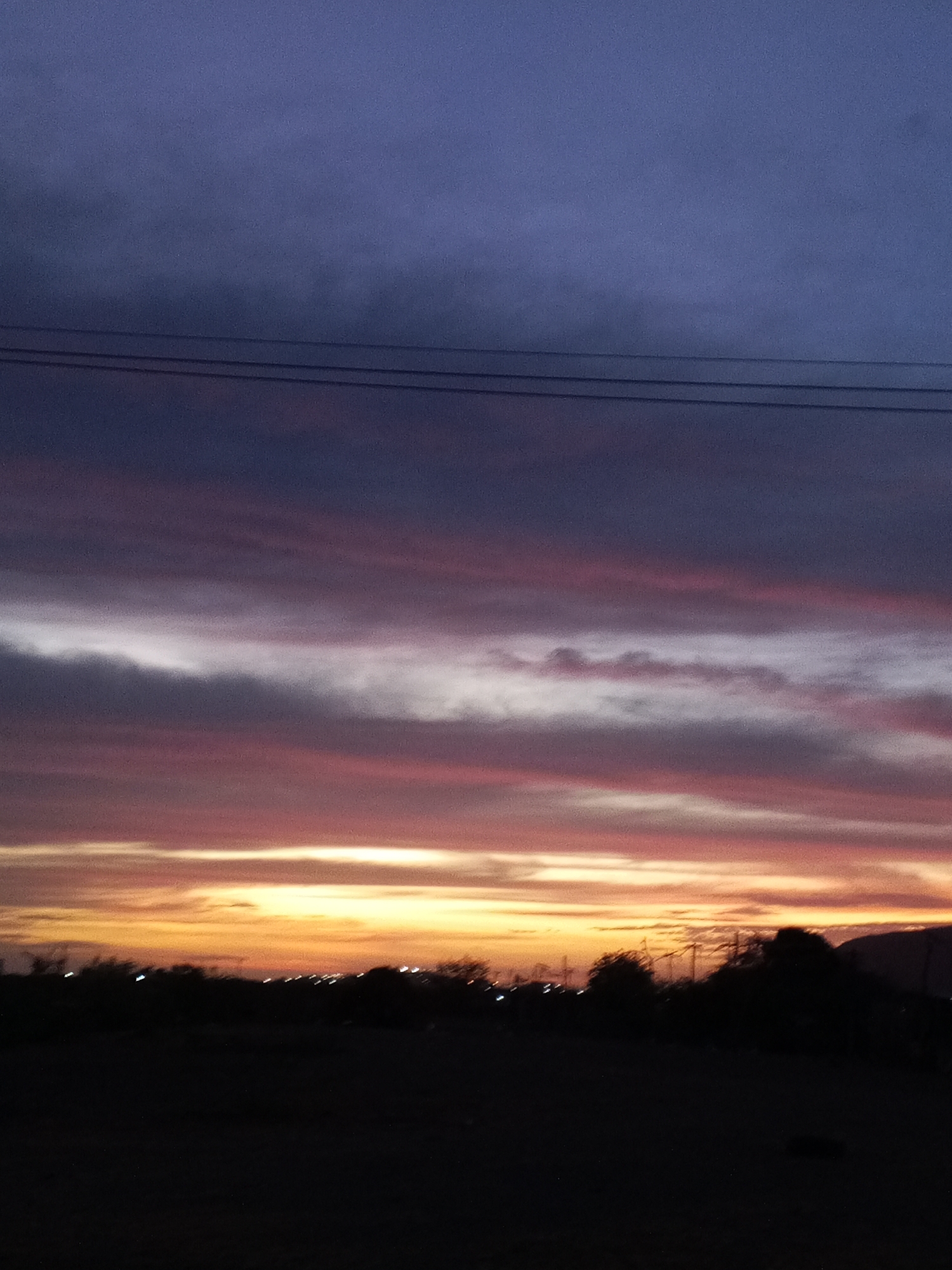 A landscape photograph depicts a sunset over Altagracia, Venezuela. The sky transitions from deep purple at the top to bands of pink, white, and orange closer to the horizon. Silhouetted against the light are trees, power lines, and a distant hill. Scattered points of light suggest the presence of the city in the background. The foreground is dark and largely obscured, indicating a field or open area.