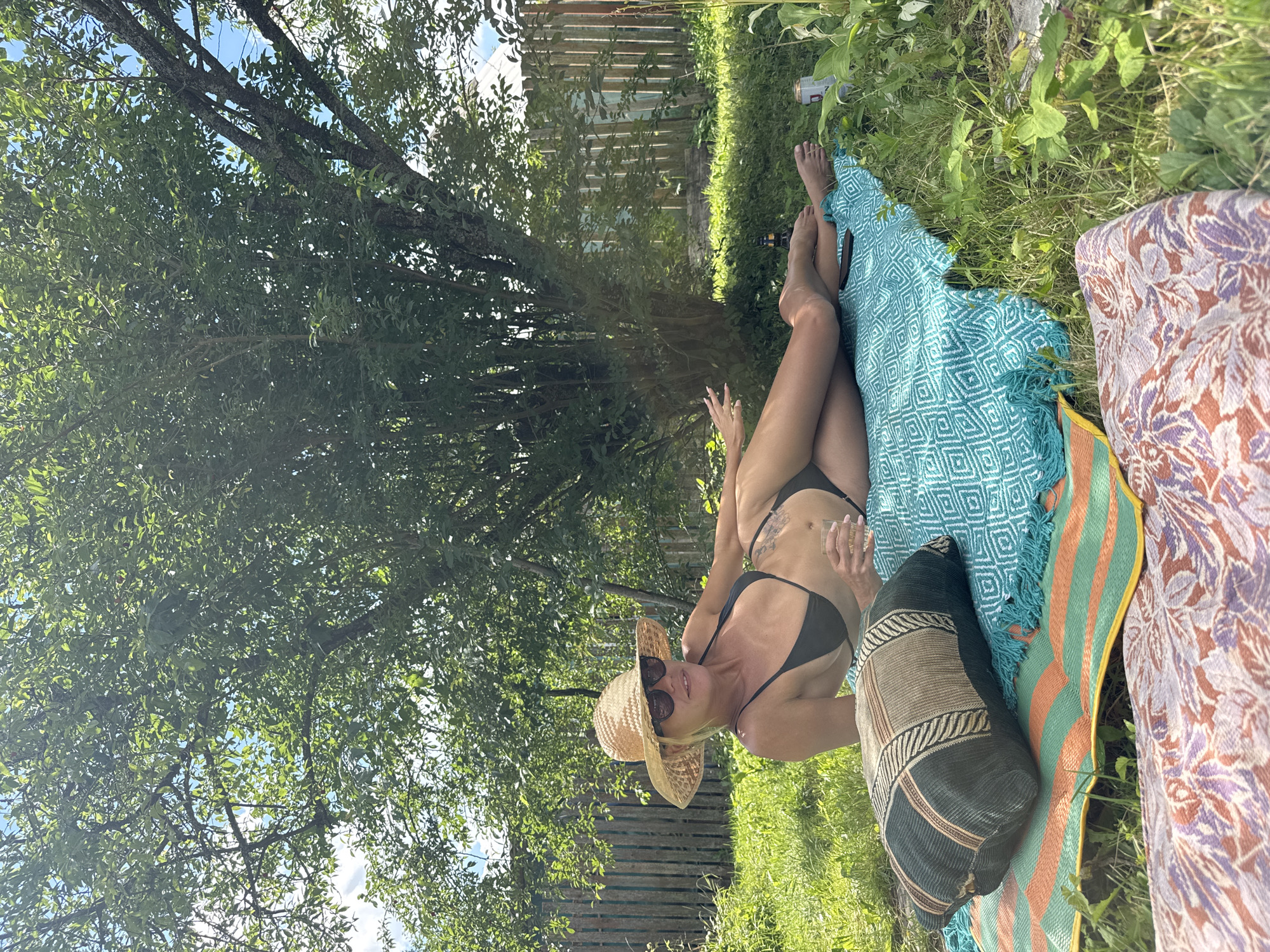 In the city of Krasnaya Presnya SNT, Russia, a woman is lying on a teal blanket in a grassy backyard on a sunny day. She is wearing a black bikini, a straw hat, and sunglasses. A pillow with a geometric pattern is placed on the blanket near her head. She holds a clear glass in her right hand. The background features a wooden fence and a large tree with green leaves. Several blankets are laid out on the grass. There are also a beer can and a bottle near the woman's feet.