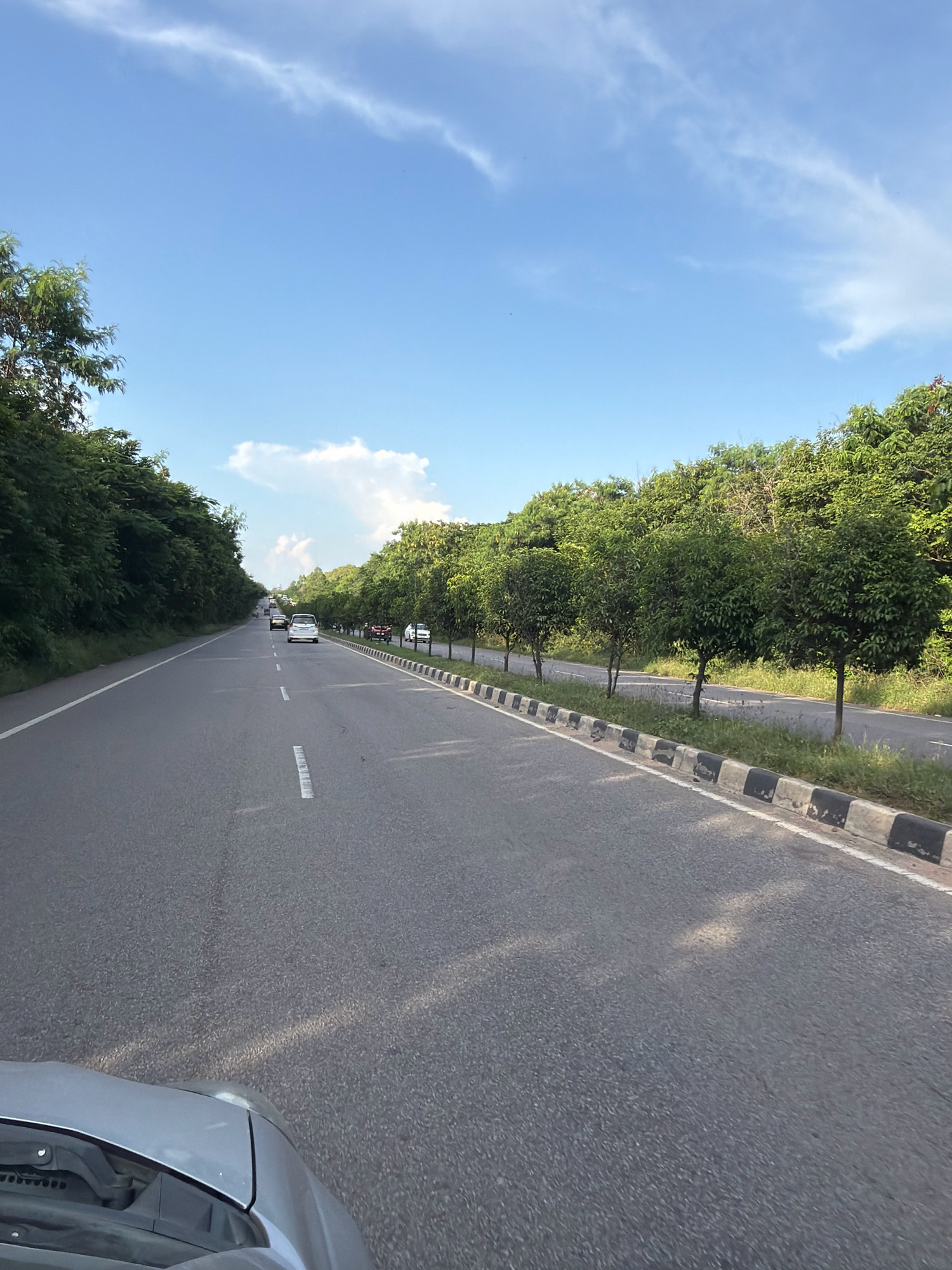 The image depicts a two-lane asphalt road lined with trees on both sides, suggesting a rural or suburban environment. The sky is blue with scattered white clouds. Several vehicles, including cars and motorcycles, are visible on the road, indicating moderate traffic. The road has white lane markings. The foreground shows the hood of a silver car, implying the photo was taken from inside a moving vehicle. The trees appear lush and green, suggesting a warm season. The setting is consistent with a road in or near Turakapally, India, a location known for its green landscapes and developing infrastructure. The image captures a snapshot of daily life on a road, with vehicles traveling and nature providing a scenic backdrop.