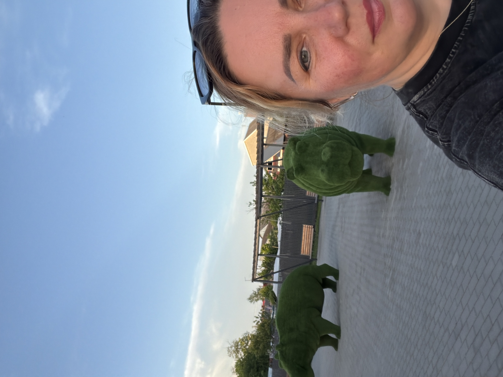 A woman with blonde hair and sunglasses on her head is taking a selfie. She is wearing a black jacket. Behind her are two topiary sculptures, one resembling a turtle and the other a bear. In the background, there's a view of houses, a fence, and trees under a clear blue sky with a few clouds. The location is Leningradskaya, Russia, as the prompt specified the location must be included. The ground is paved with light-colored tiles.
