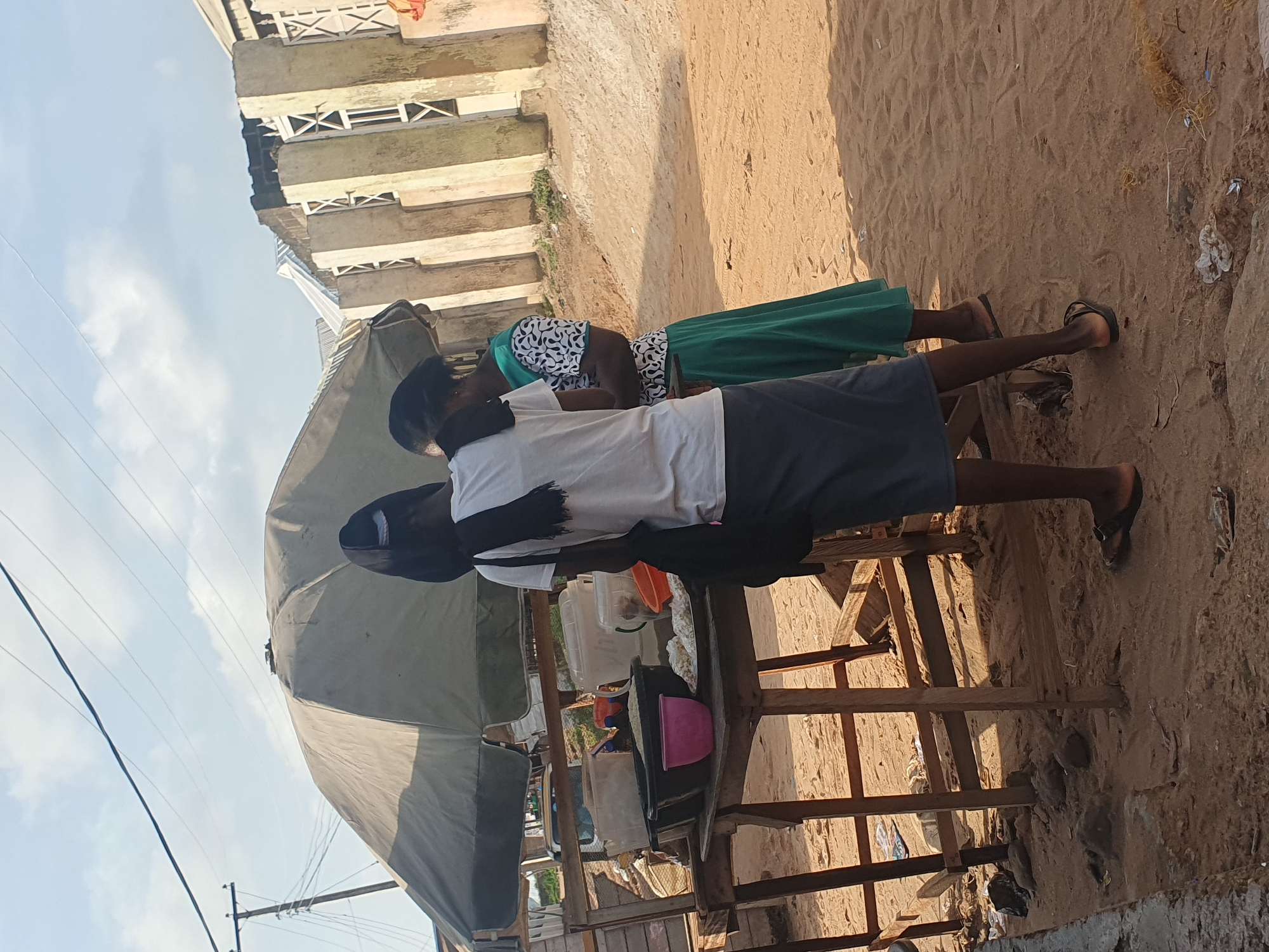The image shows two individuals near a market stall in Lokoja, Nigeria. The stall has a large, olive-colored umbrella providing shade over a wooden table laden with various containers of food items. One person, seen from the back, wears a white t-shirt and a dark skirt and has a black bag slung over their shoulder. The second person, facing slightly away from the camera, is wearing a green skirt and a patterned top. The ground is sandy, typical of an outdoor market setting. In the background, there are buildings with distinct architectural features, suggesting an urban environment. Power lines and a utility pole are visible in the sky, indicating the presence of infrastructure. The overall scene depicts a typical day at a Nigerian market.
