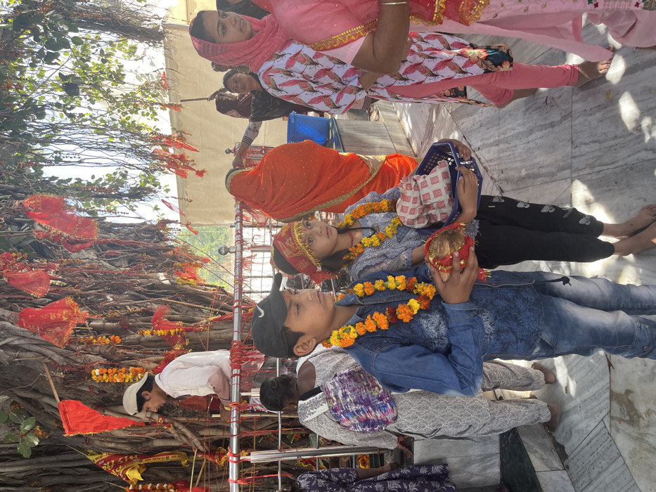 The image shows a scene at a temple in Panchkula, India, located at the coordinates 30.724 latitude and 76.859 longitude. The scene is dominated by a large tree with numerous branches and hanging vines. People are gathered around the tree, primarily women and children, some wearing traditional Indian clothing. A young boy in a blue denim shirt and a girl with a red and gold head covering are holding offerings in their hands. Other individuals are standing nearby, including a woman in a pink and white patterned dress and a woman in a bright orange sari. The background features a white tent and other trees. The ground is covered with white marble tiles. The overall atmosphere is serene and celebratory.