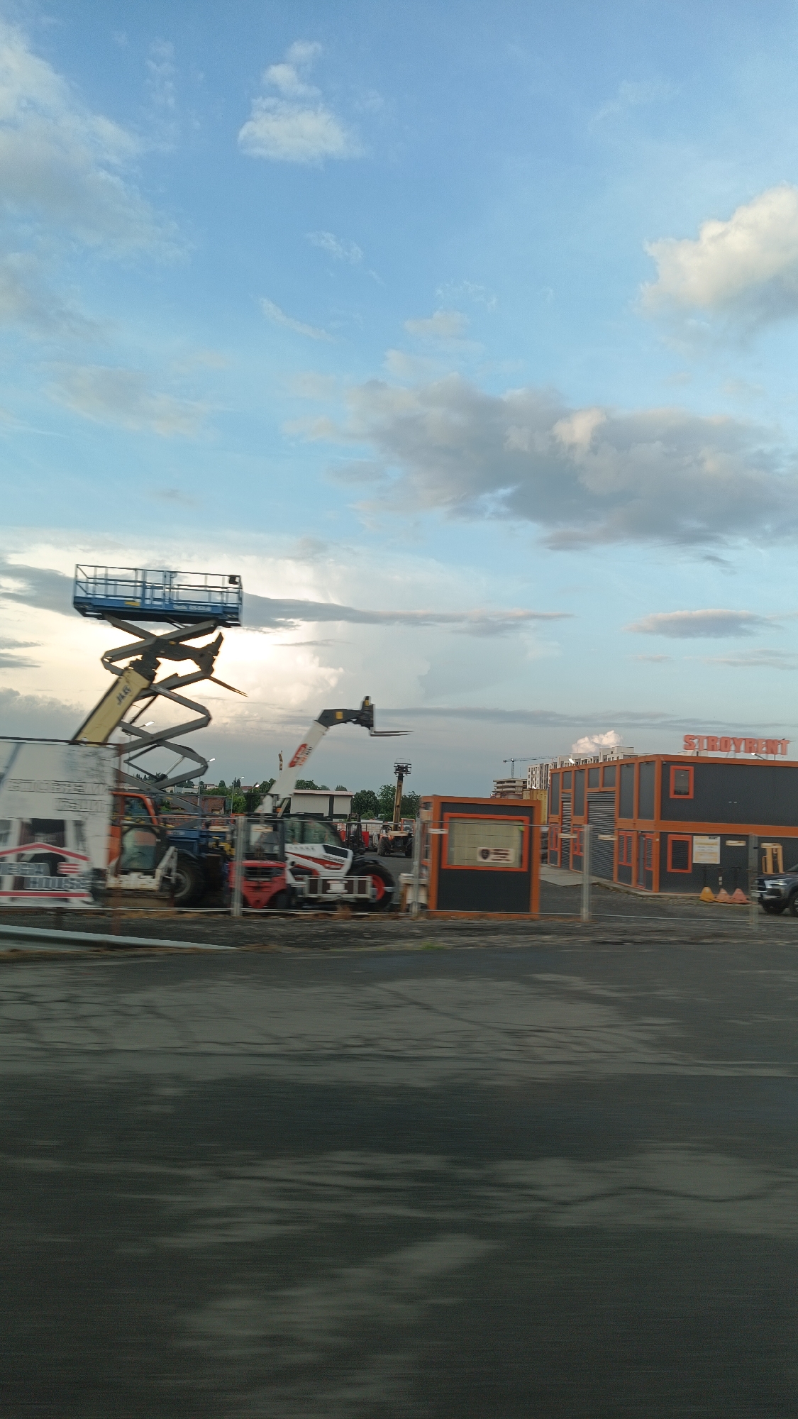 The image depicts a construction site in Burgas, Bulgaria, under a sky with patchy clouds. Prominently featured is heavy machinery, including a blue platform scissor lift extended upwards and several forklifts or similar vehicles. Behind these, a row of orange-framed, modular container units, one labeled "SIROYRENT," are visible. There is a security or information booth next to these units. The foreground shows an expanse of wet pavement or ground, giving the impression of recent rain. A fence partially obscures the view of the machinery, and an advertisement is visible on the fence. Construction appears to be ongoing as indicated by the crane in the distance.