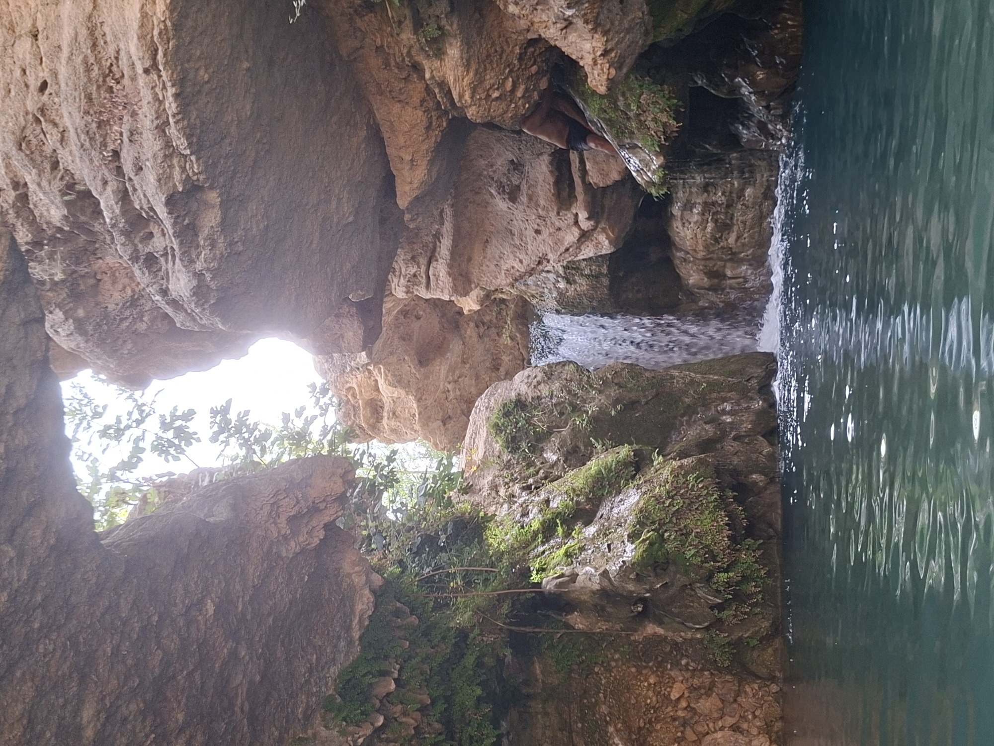 A person is partially visible, with their legs and torso obscured by rocks as they crouch or crawl within a narrow canyon. The environment is characterized by a natural pool of clear, teal water in the foreground, with a small waterfall cascading down a rocky cliff face into the pool on the left side of the frame. The canyon walls are composed of reddish-brown rock, with moss and greenery growing on some of the wetter surfaces. An archway formed by the rock overhang at the top of the frame allows sunlight to illuminate the scene. The location is described as Unknown, Spain.