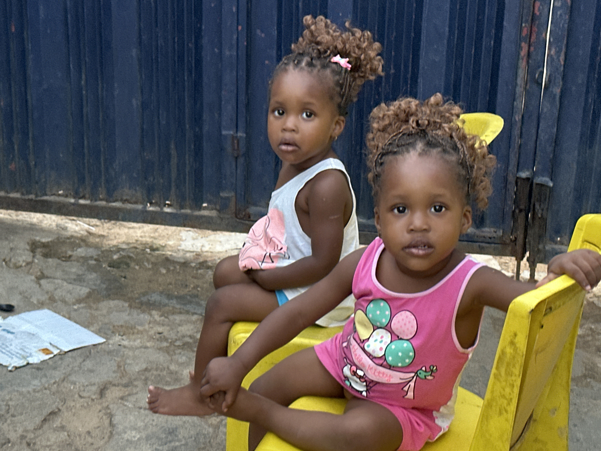 Two young girls are seated outdoors in Narayi, Nigeria, looking directly at the camera. The setting features a prominent dark blue corrugated metal fence or wall in the background, suggesting an informal urban or semi-urban environment. The ground is a mix of concrete and dirt, with a piece of crumpled paper or cardboard visible on the left. The scene is brightly lit, indicating daytime.
The girl on the left, slightly behind the other, wears a white sleeveless top with a light pink and white patterned design, and blue shorts are visible underneath. Her dark, curly hair is styled in braids gathered into a high puff, secured with a small pink hair tie. She has a neutral, somewhat curious expression.
The girl in the foreground is seated on a bright yellow plastic child's chair, which shows signs of wear. She is dressed in a matching pink sleeveless top and shorts set. Her top features a "Hello Kitty" design with various colored balloons (pink, light blue, white, and green with polka dots) and a small airplane motif. Her hair is styled similarly to the other girl's, with braids gathered into a high puff, adorned with a yellow hair tie or bow. She also has a neutral to slightly pensive expression. Her bare feet are visible, resting on the ground or the chair. The girls appear to be siblings or close companions.