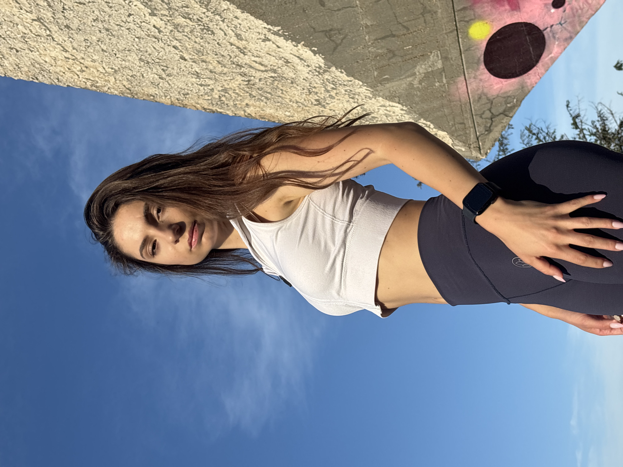 A woman stands outdoors against a clear blue sky, with a rough concrete structure to her right. She is wearing a white cropped zip-up top and dark leggings, and a smartwatch on her left wrist. Her long brown hair is loose. The concrete structure has graffiti on it, including pink and yellow circles. The scene is likely in a park or an urban area with public art. It is plausible this is in the Sea Garden of Varna, Bulgaria, given the combination of open spaces, concrete structures, and potential for public art installations.
