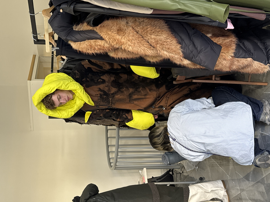 An indoor scene located in Milan, Italy, depicts a fashion preparation area. Central to the frame, a young male with curly light brown hair stands, dressed in an oversized, dark brown and black tie-dyed or bleached hooded garment, potentially a jumpsuit. The garment features a large, puffy, neon yellow hood enveloping his head and neck, and matching puffy neon yellow sleeve cuffs. He looks downward and to his right. In front of him, a second individual, with short light brown hair, kneels on a grey tiled floor. This person wears a light blue and white striped long-sleeved shirt, dark blue jeans, and grey sneakers, with a dark cord hanging over their right shoulder. They are actively adjusting the lower leg area of the standing individual's attire. To the far right, a clothing rack holds multiple garments, including a black puffer jacket detailed with a substantial brown faux fur panel. A dark wooden chair is partially visible behind the kneeling person. On the left side of the frame, a partial view of a third person in dark clothing is visible. A white table with small items, including a pink object, and a grey cylindrical container are on the floor to the left, near a curved vertical metal railing. The environment features plain off-white walls.