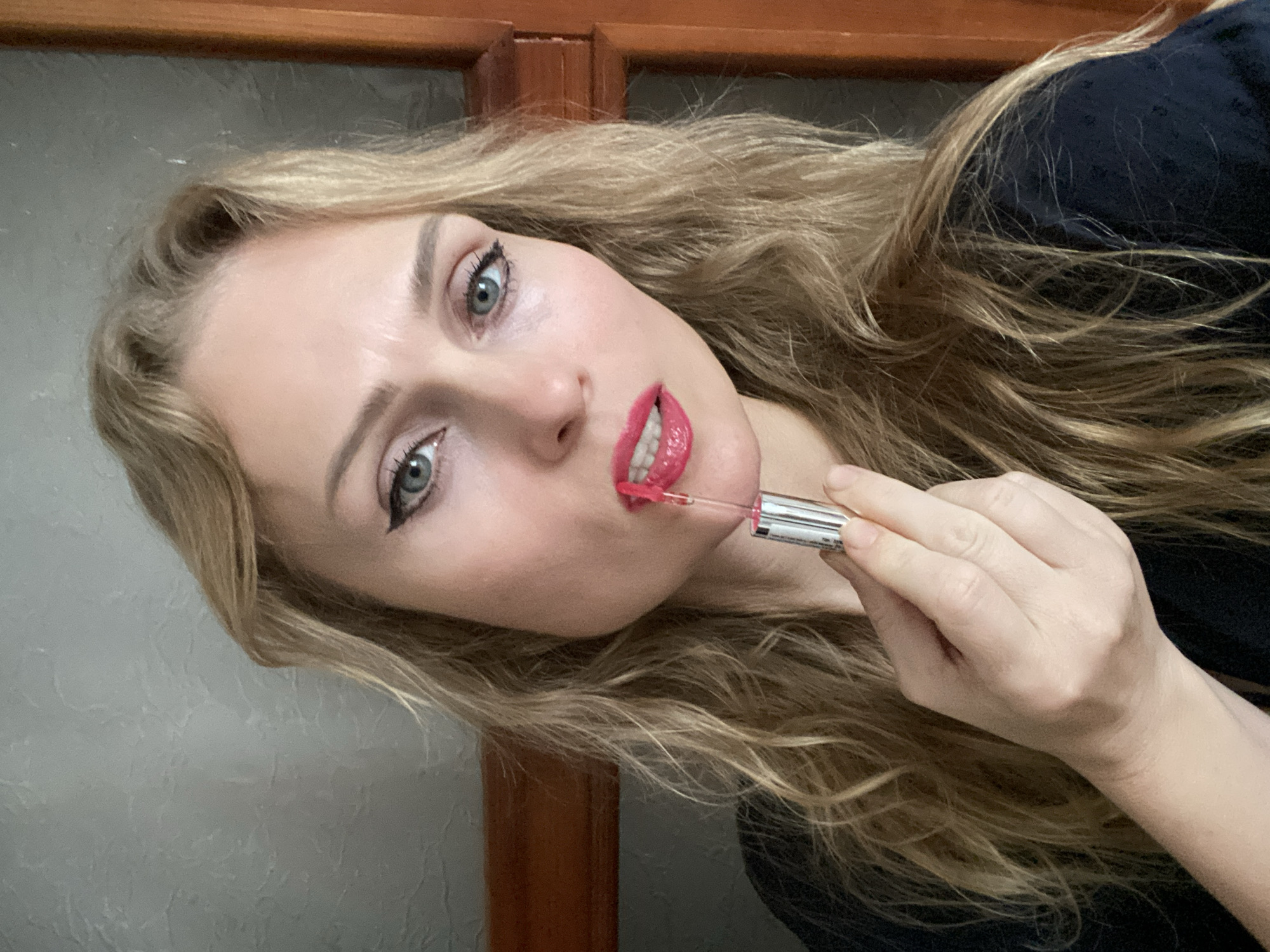 In an indoor setting, a woman with blonde, wavy hair and blue eyes is applying bright pink lipstick. She is holding the lipstick applicator with her right hand, and her lips are parted as she applies the product. She is wearing a dark-colored top. The background appears to be a wall with a textured surface, and a wooden doorframe is visible on the right side of the image.