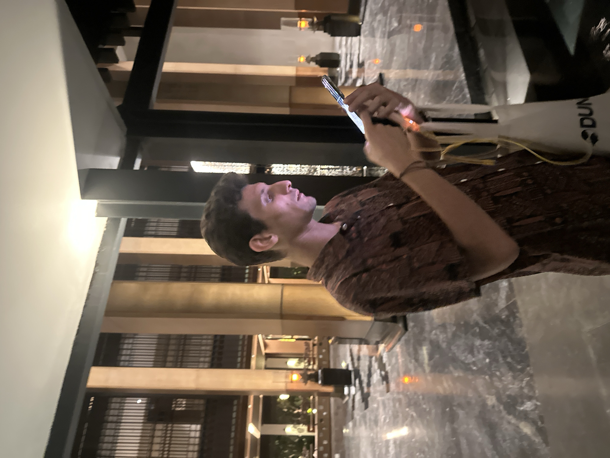In the image, a man with short, dark, curly hair is looking at his phone. He is wearing a brown shirt with a pattern. He is standing in what appears to be a modern indoor space with marble flooring. The background features architectural elements such as wooden beams and railings, along with some lighting. A white bag with the text "DUN..." is visible near his right arm. The lighting suggests it is either evening or an indoor setting with artificial lights. The man's expression seems focused on his phone. The scene is static, with the man engaged in using his device. There are no clear indicators of weather or time of day beyond the artificial lighting. The overall impression is one of a casual moment in an indoor public or private space.