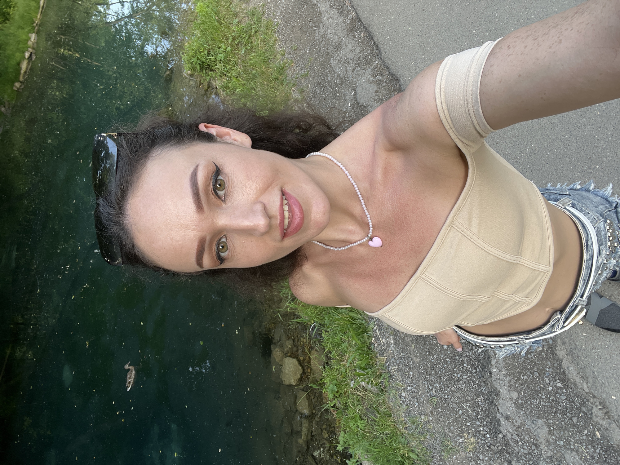 In a medium shot, a woman with long, dark, wavy hair and light green eyes is taking a selfie by a body of water. She is wearing a light beige, off-the-shoulder top and distressed denim shorts. A pearl necklace with a pink heart pendant adorns her neck. She has on winged eyeliner and lip gloss. Sunglasses are perched on her head. Behind her, the water is a deep blue-green, with a duck swimming in the distance. The bank of the water is lined with rocks and green grass. A paved path is visible to her right. The scene is outdoors, with the lighting suggesting it is daytime. The location is identified as being in the city of Kazan, Russia.