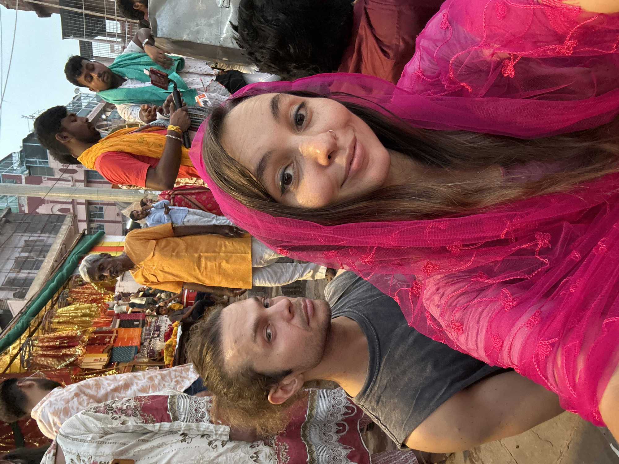 The image is a selfie taken by a woman wearing a pink shawl or dupatta. A man is next to her. Both of them are facing the camera. The background consists of other people, clothing, and buildings. Several men are standing in the background, and the one closest to the top left is using a smartphone. The scene appears to be a busy market or public gathering place in the city of Varanasi, India. The lighting suggests it is daytime. There is a shop selling colorful fabrics in the left of the image.