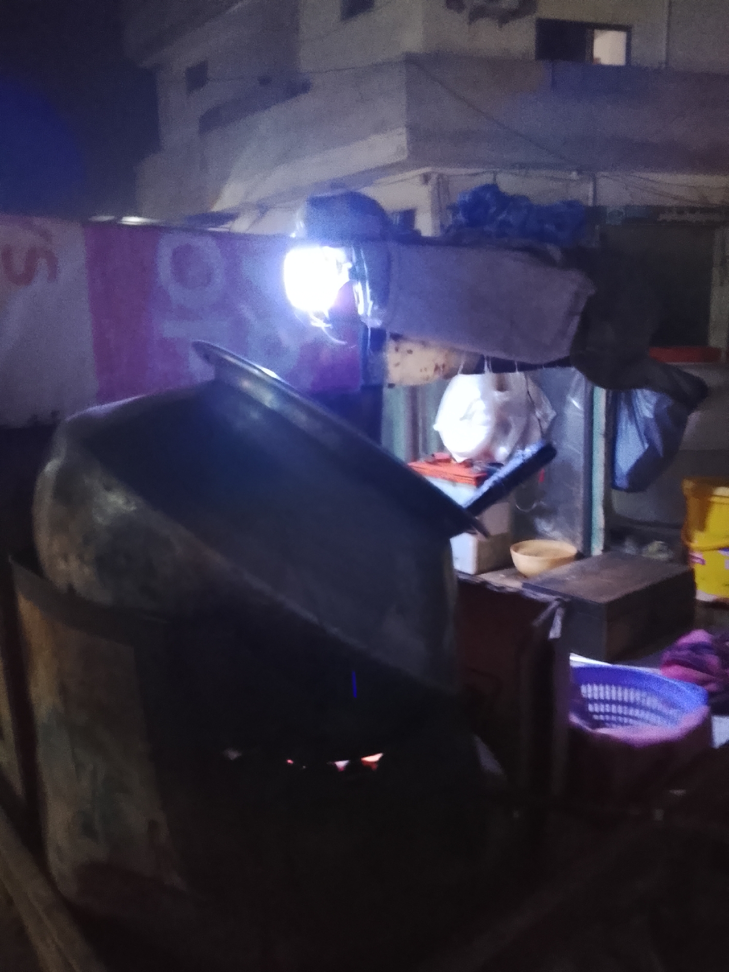 The image captures a nighttime scene of a street vendor setup in what appears to be Nasheman Colony, Pakistan. No people are visible in the frame.
The central focus is a large, metallic cooking pot or wok, angled downwards and resting on a dark, utilitarian stand. Below the pot, an orange glow suggests a heat source, likely an open flame or embers, indicating cooking activity. A very bright, exposed light bulb or LED fixture hangs above and to the right of the pot, intensely illuminating the immediate area and creating strong highlights and shadows.
Behind the pot, various items are stored and arranged. Several white plastic bags hang, one appearing to contain something spherical. A small, light-colored bowl (possibly yellow or beige) sits on a wooden surface. Other dark boxes and containers are visible. To the far right, yellow plastic containers are stacked, and a blue plastic basket with a distinct grid pattern is in the lower right foreground. Dark blue bundles or tarps are piled on top of the stall structure, suggesting coverings or stored goods.
In the background, parts of a building with windows are dimly visible against the dark night sky. The left side of the image shows a banner or sign, partially obscured, with visible white letters including a capital "S" and "TO" on a reddish-pink background. The overall impression is that of a functional, albeit rustic, outdoor food preparation area operating after dark.