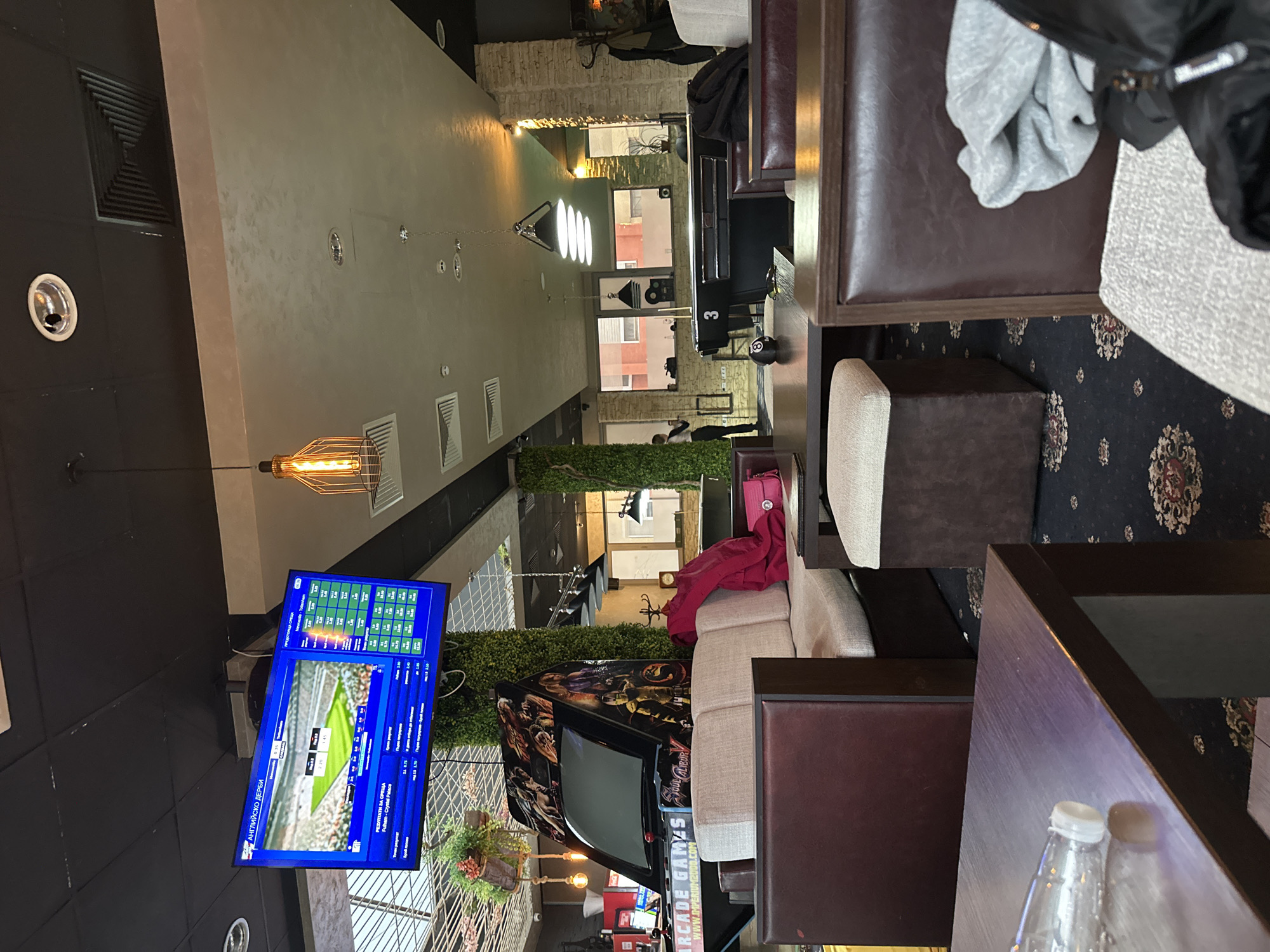 Here's a detailed description of the image:
The image appears to be taken in an indoor space, likely a bar or lounge, in Plovdiv, Bulgaria. There is a television on the left-hand side displaying a sports game, which seems to be a soccer match (Fulham-Crystal Palace). Above the game information, there are betting statistics.
In the foreground, there is lounge furniture consisting of chairs and tables. There is a bottle of water on the table.
Beyond the seating area, the space extends further inside. There's a second gaming machine.
The interior design includes decorative lighting fixtures hanging from the ceiling and greenery along the walls. The walls appear to be textured, and there are ventilation ducts visible. The carpet has an ornate floral pattern.
The scene shows a combination of a relaxing lounge area and elements of a sports bar with arcade games.
