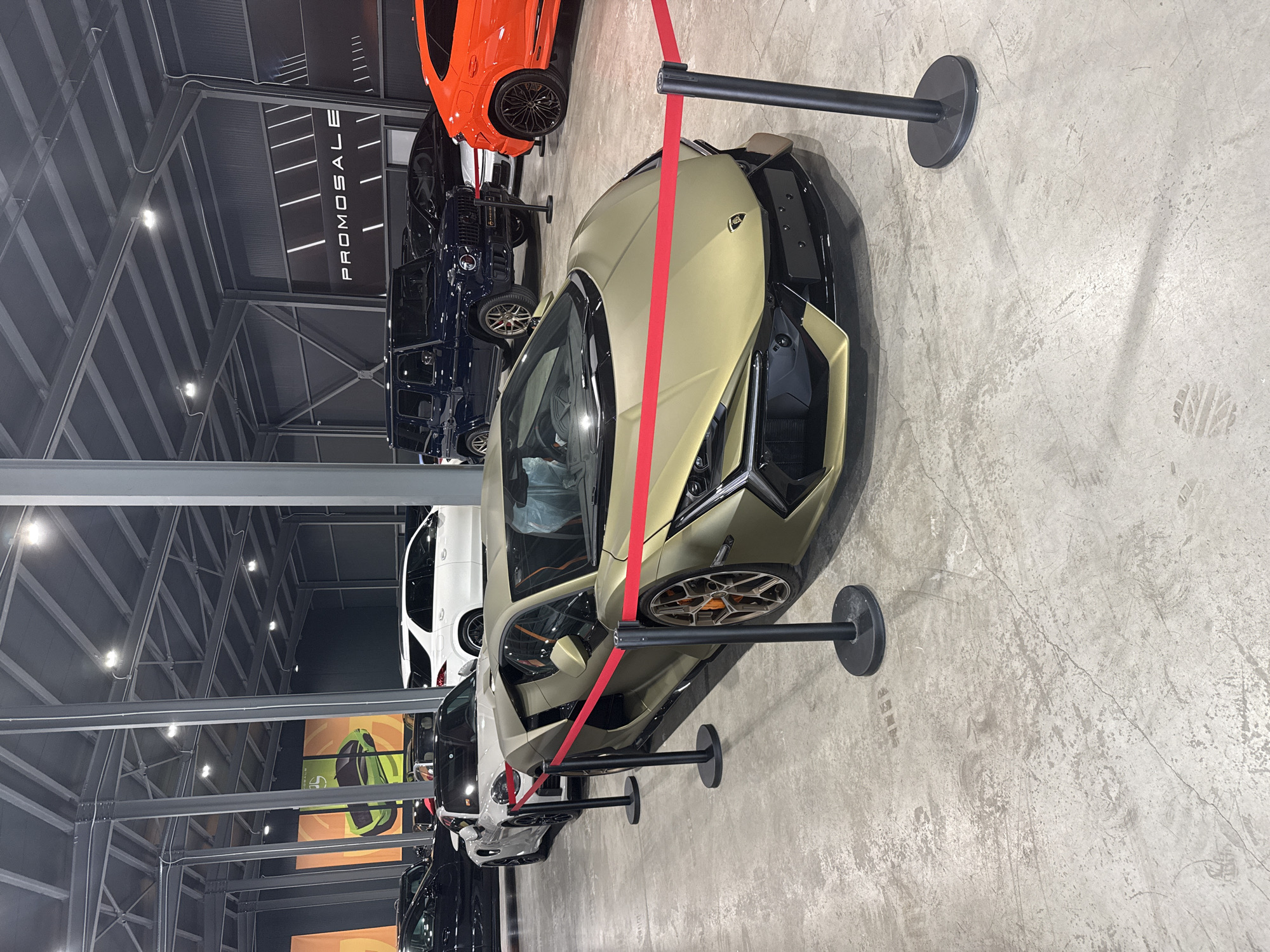 The image shows a showroom with several luxury cars on display, likely in Plovdiv, Bulgaria. There is a gold-colored Lamborghini at the forefront, separated from the other cars by a red rope barrier. Behind the Lamborghini, there are several other vehicles, including a blue SUV and an orange car. The building's interior features a high, industrial-style ceiling with metal beams. The floor is concrete. A sign with the word "PROMOSALE" is visible in the background. There are no people visible in the immediate scene.