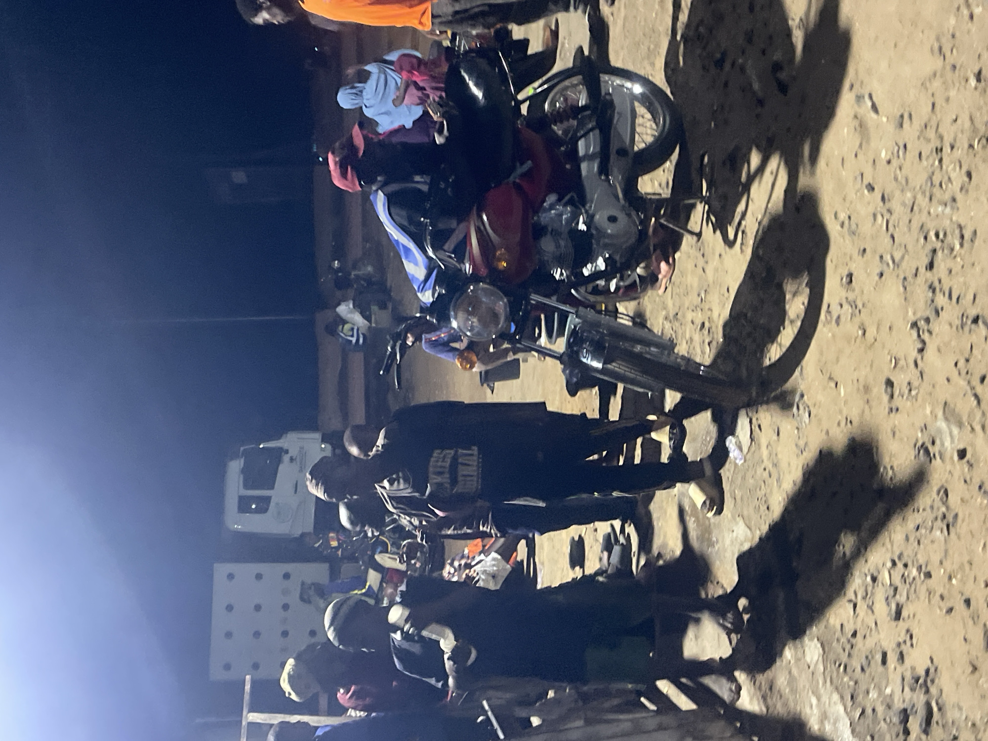 In Lokoja, Nigeria, at night, a group of people are gathered around a motorcycle in an outdoor setting. The ground is covered with dirt and small stones, and there are prominent shadows cast on the ground, indicating artificial lighting.
In the foreground, a red motorcycle is parked, with its kickstand down. A person wearing a red cap and a blue and white jacket is seated on the motorcycle. To the left of the motorcycle, two individuals are standing. The person on the far left wears a dark t-shirt and green shorts, holding a white object. Next to them, another person is dressed in a dark t-shirt with "Dickies Original" printed on it and dark pants.
In the background, a white truck with a large cargo box is visible. The truck has "DANGOTE" written on its side. Several other people are scattered throughout the scene, some standing and some appearing to be seated. One person in the mid-ground wears a light blue head covering and pink clothing. The overall atmosphere suggests a busy gathering or temporary stop, possibly for a meal or rest.