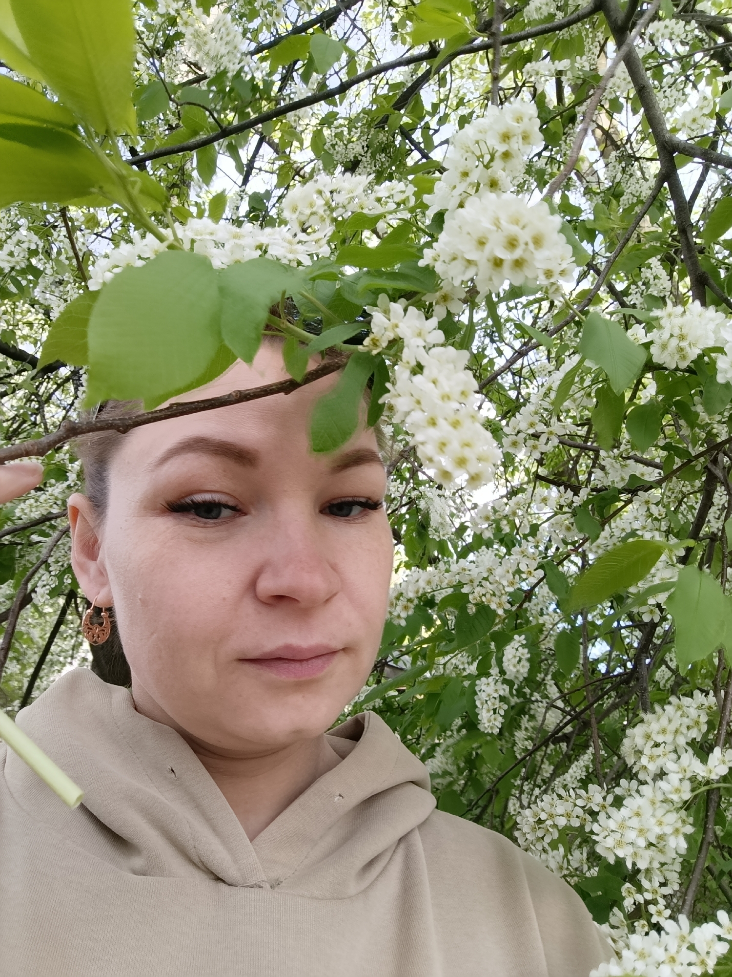 A woman is standing beneath a flowering tree in Chaikovsky, Russia. The tree is laden with white blossoms and green leaves. The woman is wearing a beige hooded sweatshirt and ornate gold earrings. A green straw is visible near her shoulder. Her gaze is directed slightly downward.