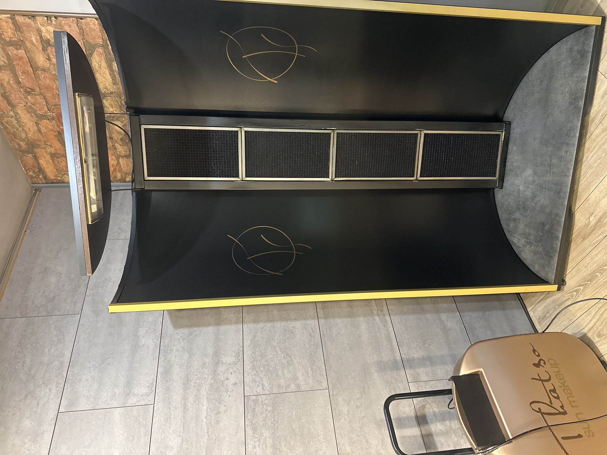 The image shows a tanning bed in a room with gray tile flooring. The tanning bed is primarily black with gold trim and features a design on the upper and lower portions. Five square vents are visible in the center section of the bed. The bed is open, with one side panel standing upright. Adjacent to the tanning bed, a chair with a gold seat labeled "Ratso sun makeup" is visible. The background includes a wall with exposed brick. The location appears to be indoors, potentially a tanning salon. While no definitive information about Riga, Latvia is available from the image, it is possible that the tanning salon is located there if that is the intent of the prompt.