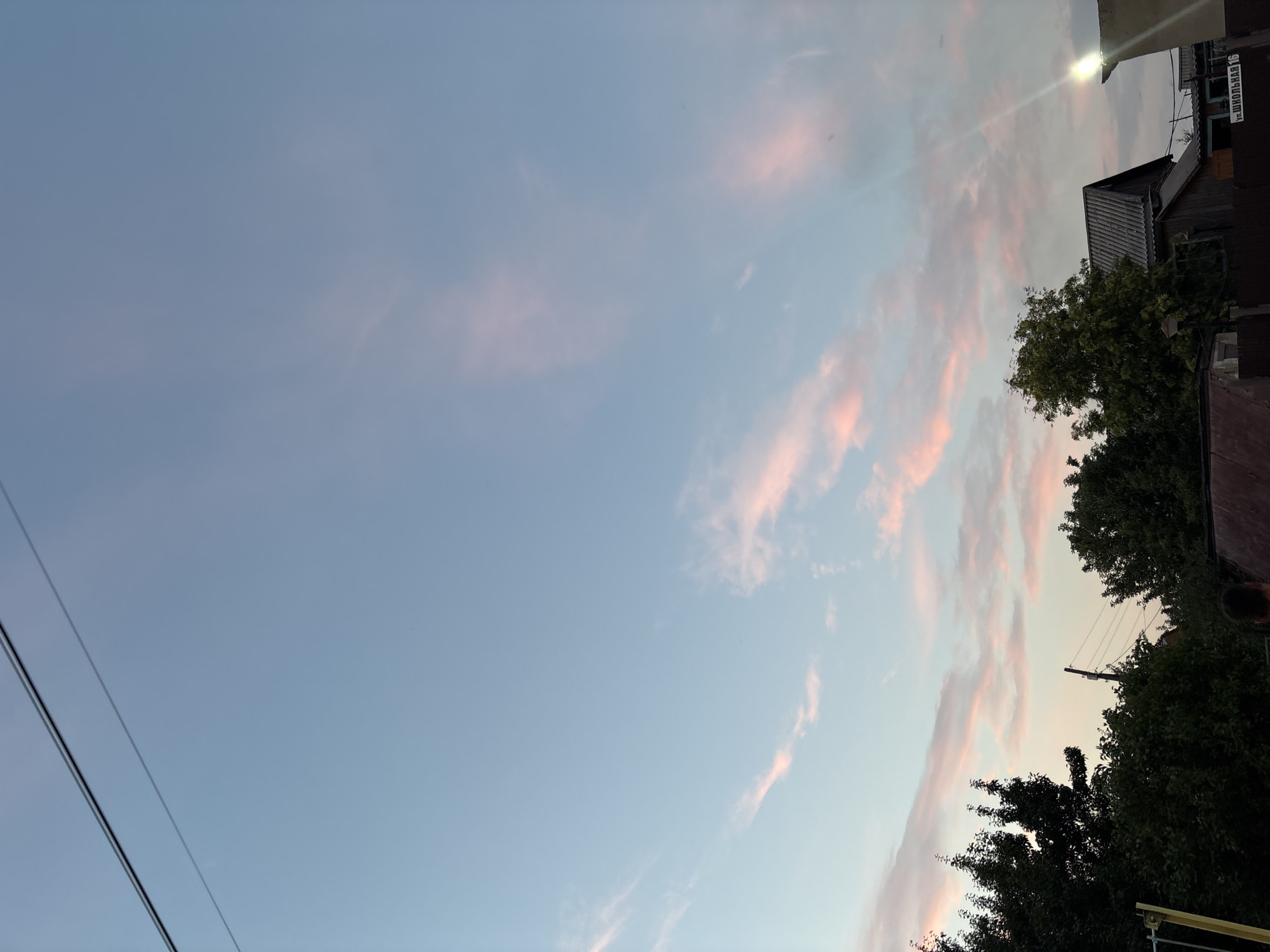 This is a photograph taken in Vishnevka, Russia, depicting a rural landscape at dusk. The sky dominates the upper two-thirds of the frame, transitioning from a pale blue at the top to lighter hues near the horizon, interspersed with wispy clouds tinged with pink and orange, indicative of sunset or sunrise. Two dark wires stretch diagonally across the upper left portion of the sky.
In the lower portion of the image, a line of dense green trees forms a backdrop, with their foliage silhouetted against the sky. To the right of the trees, several buildings are visible. A grey, pitched-roof house with a weathered exterior stands prominently. Adjacent to it, to the right, is a smaller building with a light fixture shining brightly. A dark fence runs across the bottom of the image, separating the foreground from the structures and trees. Partially visible at the bottom center of the image is the back of a person. The overall impression is one of a quiet, end-of-day rural scene.