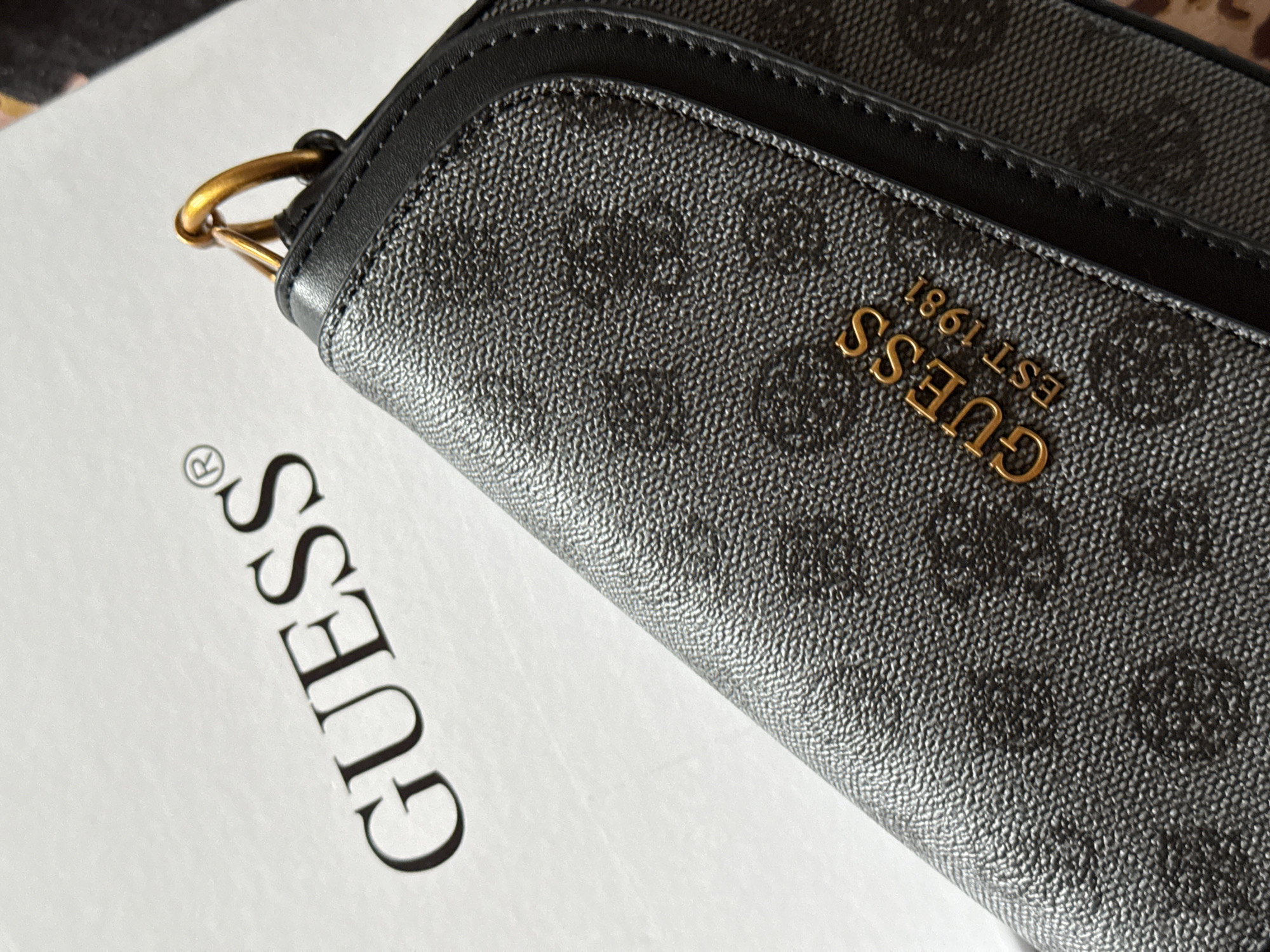 The image displays a close-up, angled shot of a black Guess wallet with a textured, monogrammed pattern. A gold-toned metal ring is attached to the wallet's corner. To the left of the wallet, the word "GUESS®" is printed in large, black, sans-serif font on a white surface, likely a box or packaging. The lighting suggests an indoor setting, possibly a retail or home environment. No people, weather, or time of day cues are visible. The wallet itself shows the "GUESS EST. 1981" logo in a gold, embossed font.
