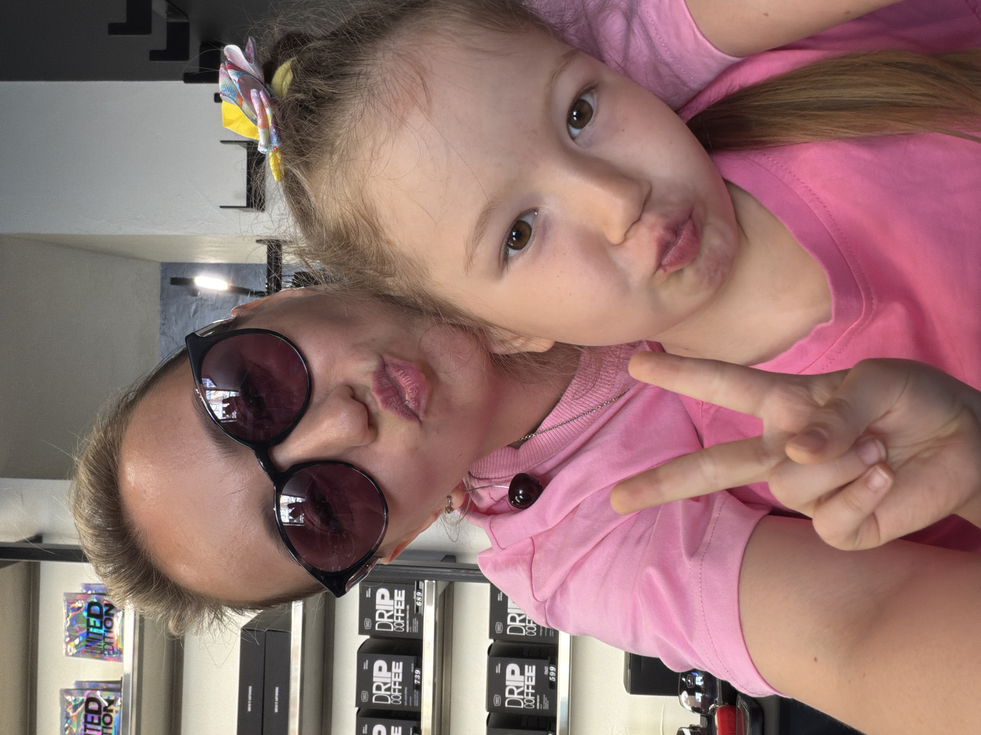 Here is a forensic description of the image:
**Scene:** An indoor setting, possibly a shop, in Izhevsk, Russia. Evidence of this location is present through the Cyrillic text on product packaging.
**People:** Two individuals, a young girl and an adult female, are posing for a selfie. Both are fair-skinned and wearing pink tops. The girl has her hair tied back with a colorful hair accessory. The adult female is wearing sunglasses and a necklace with a dark bead pendant.
**Actions/Interactions:** Both the girl and the woman are puckering their lips in a kissing gesture. The woman is holding up a two-finger peace sign.
**Objects:** A shelf behind the woman contains product packaging labeled "DRIP COFFEE" and "LIMITED EDITION". Other possible store items are visible in the background.
**Environment:** The lighting appears to be from above and slightly to the left. The background is blurred and difficult to fully discern, but seems to be a retail environment.