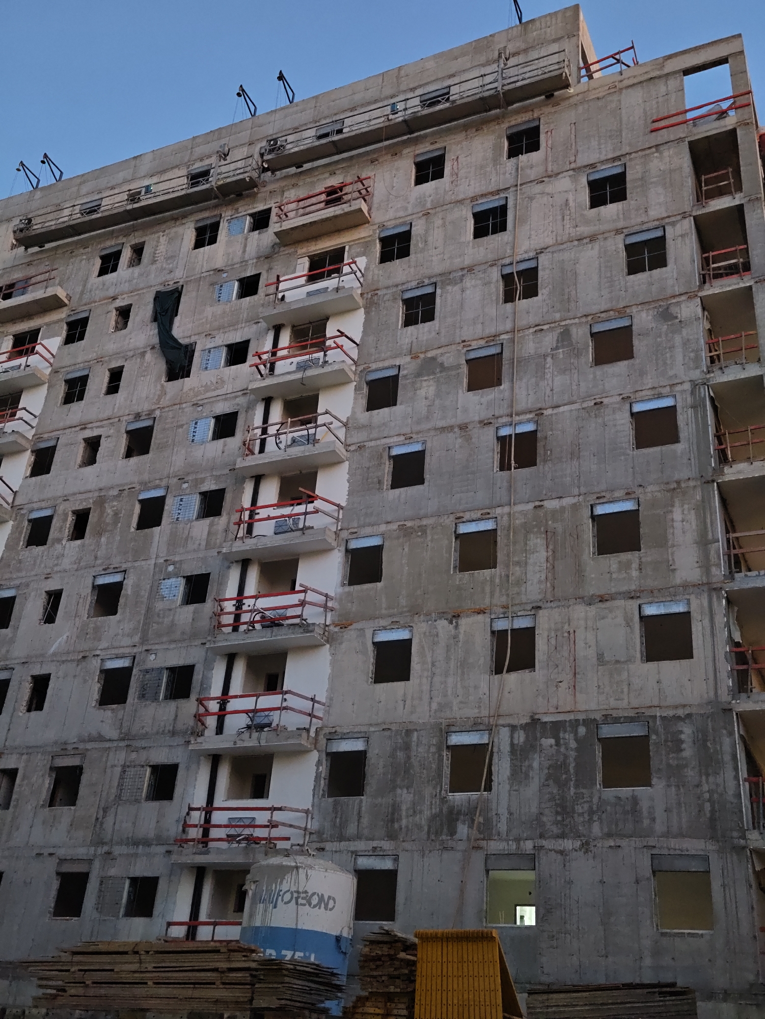 The image shows the exterior of a multi-story unfinished concrete building, likely under construction, against a clear blue sky. The building has numerous rectangular openings that appear to be window frames, some with translucent grey shutters. Several balconies are visible, each equipped with red railings and wooden supports. Scaffolding or suspended work platforms are attached to the upper levels of the building, with cables extending upwards to unseen structures. A dark green tarp hangs from one of the upper floors. In the foreground, stacks of wooden planks are visible, along with a large cylindrical white and blue object with the partial text "ALFOBO" and "75 L" written on it. The architectural style and the presence of construction materials suggest an urban or suburban setting. The building appears to be located in Petah Tikva, Israel, based on the presence of a cylindrical object with writing that likely indicates a locally sourced material or equipment. No people are visible in the image. The overall impression is one of ongoing construction in an Israeli city.