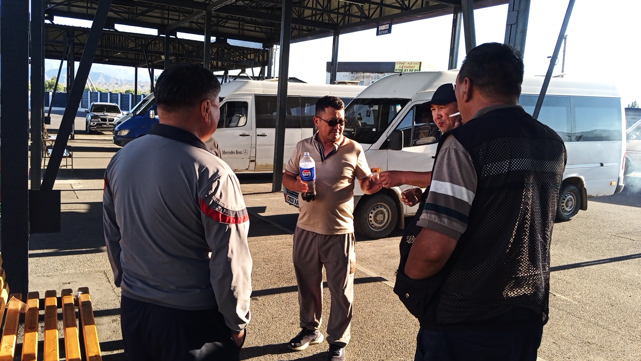 In Balykchy, Kyrgyzstan, four men stand on asphalt near a covered bus station. The man on the left, viewed from behind, wears a grey long-sleeved shirt with red and black accents on the shoulders and cuffs, and dark pants. The man in the center, facing right, wears a light brown polo shirt, grey pants, and sunglasses, holding a bottle of Pepsi. He is interacting with the man to his right, who is also facing right, wearing a grey t-shirt with horizontal stripes on the sleeves and a black mesh vest. The fourth man, on the far right, is partially visible and appears to be wearing a grey t-shirt with stripes and dark pants. In the background, white minibusses and a dark blue SUV are visible, suggesting a transportation hub. The environment is sunny with shadows cast from the covered structure overhead.